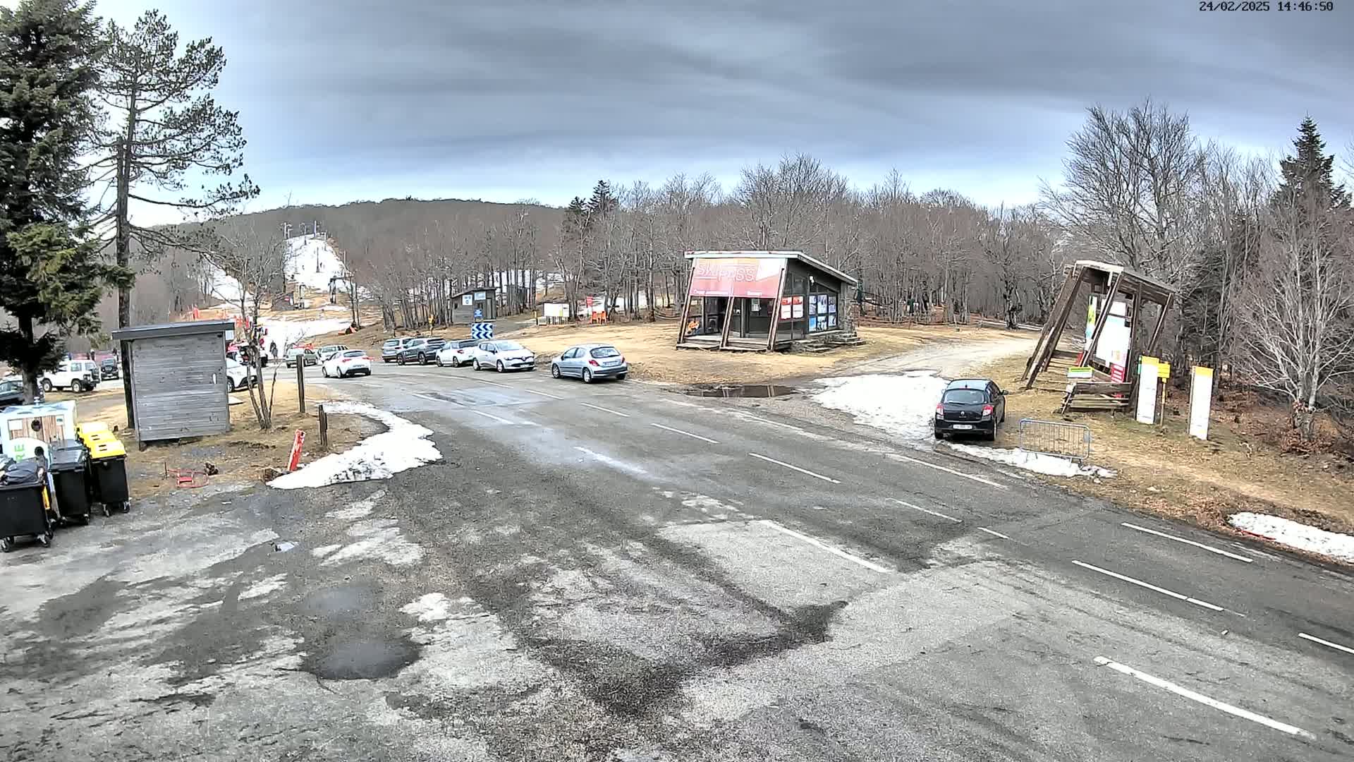 A paved road with several cars parked near a ski resort building under a cloudy sky, with patches of snow and bare trees visible.