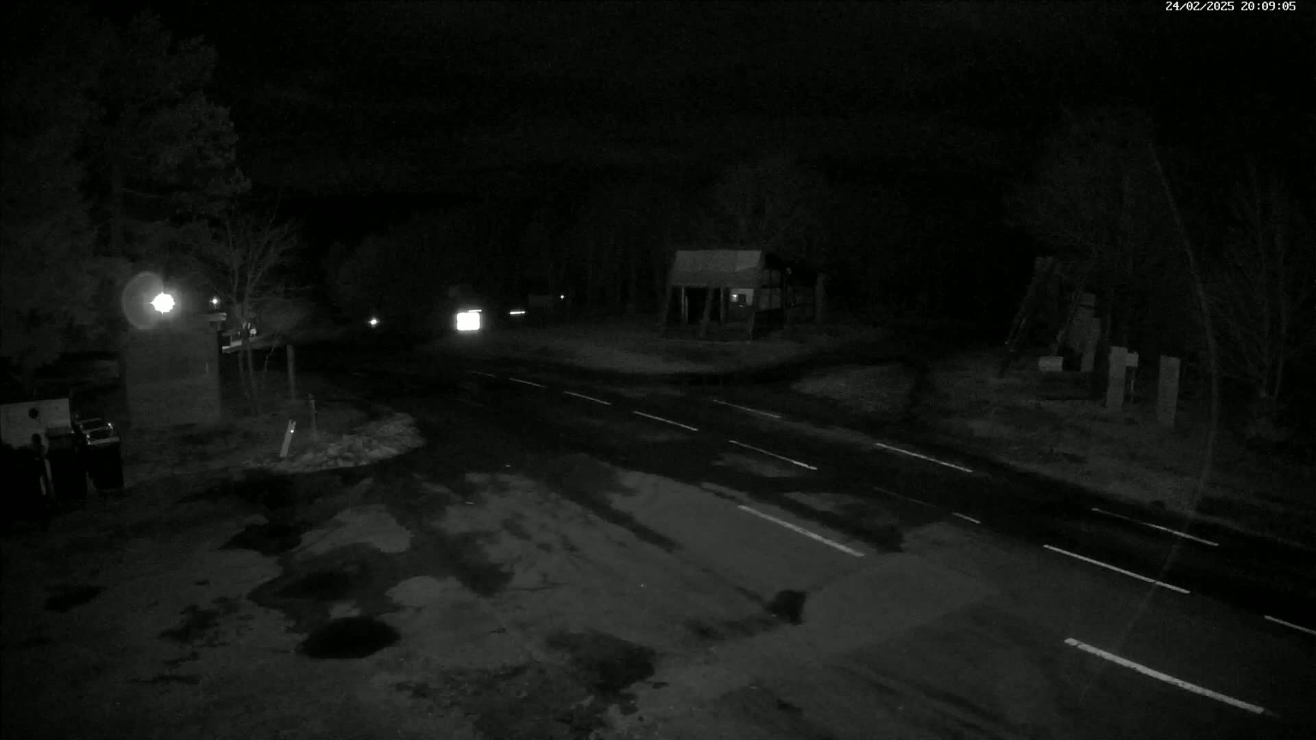 A dark, wet road intersection at night, bordered by trees and a few structures, with a vehicle visible in the distance.