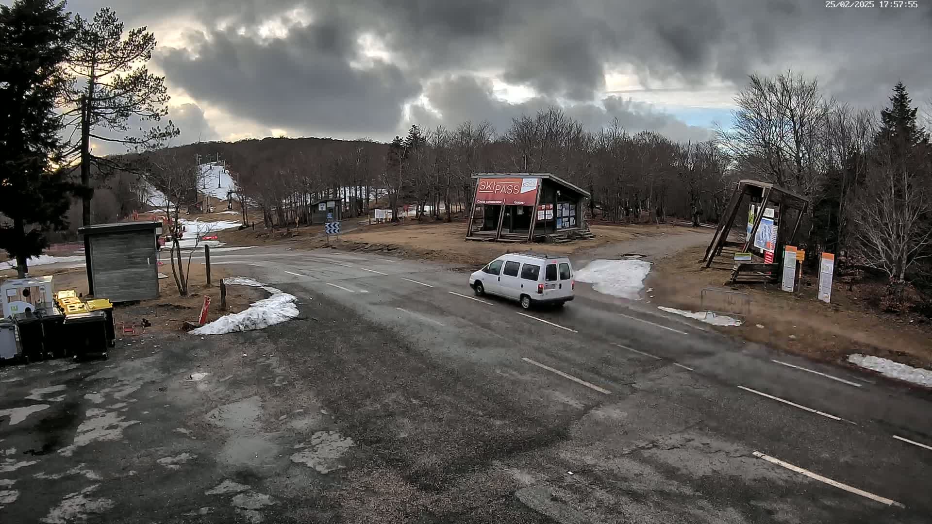 A white van drives on a road near a ski resort under a cloudy sky with patches of snow on the ground.
