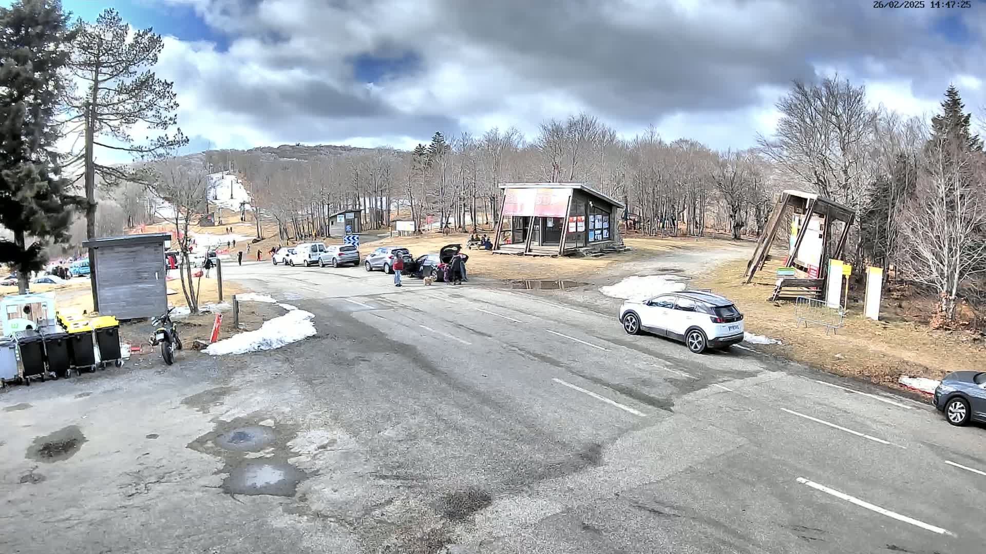 A parking lot and roadway are situated in a wooded area with a partly cloudy sky, some snow on the ground, and several cars and people present.