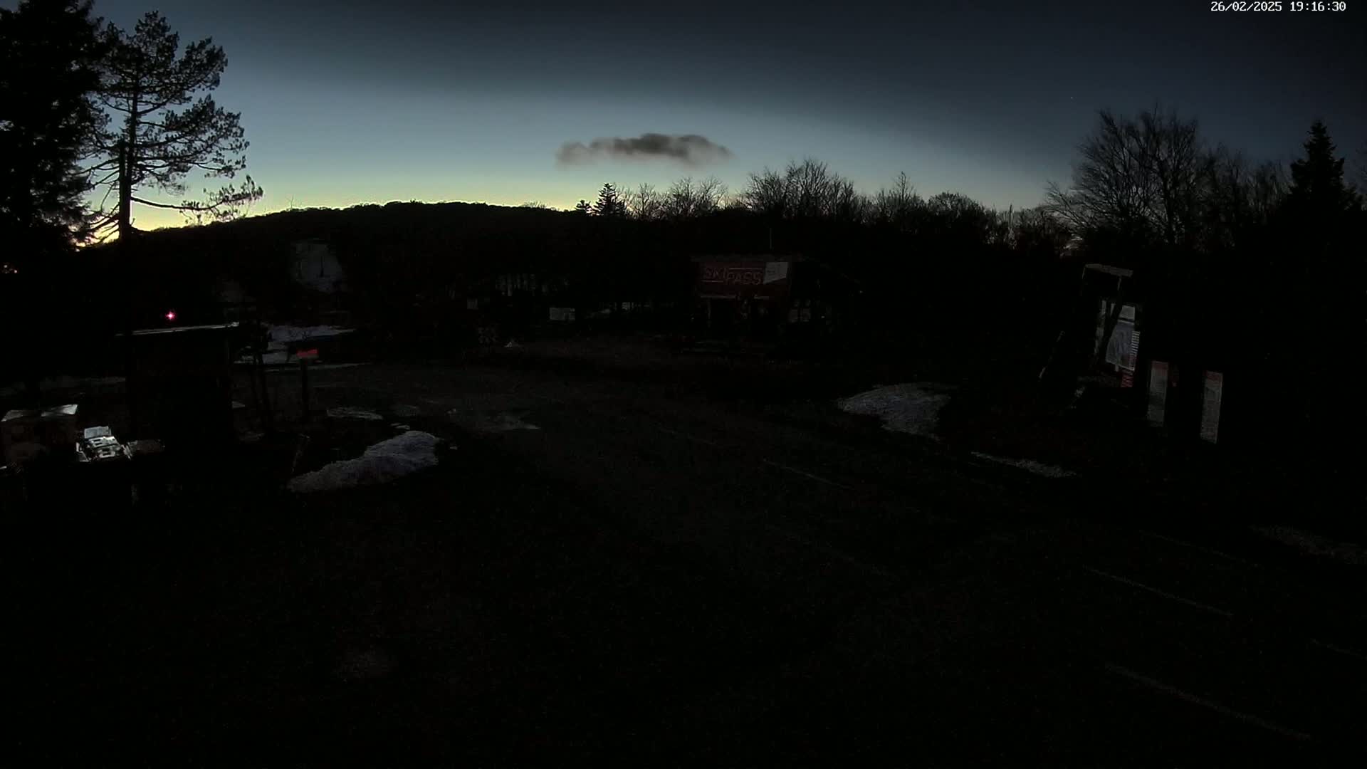 A dark, nighttime scene shows a road with a few buildings and trees under a mostly clear, twilight sky with a single cloud.