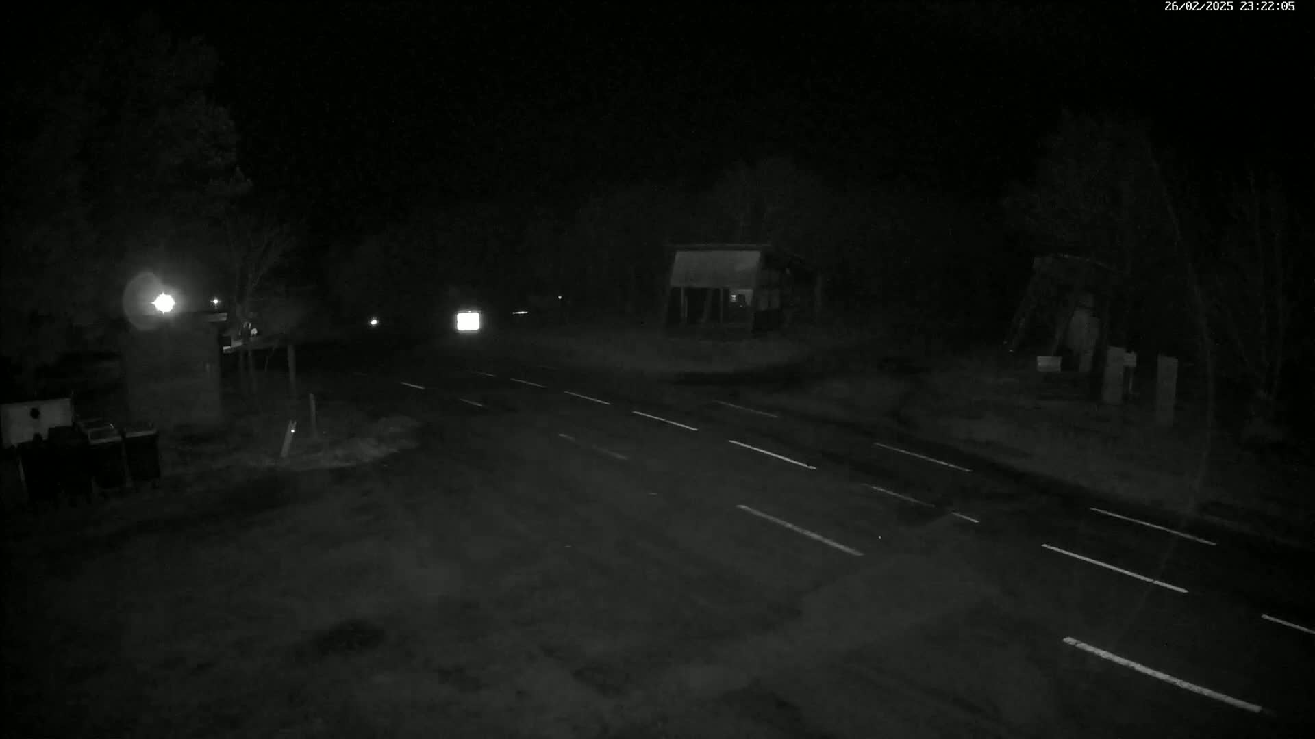 A dark, nighttime scene shows a wet, empty two-lane road with faint white lane markings, flanked by sparsely lit trees and structures.
