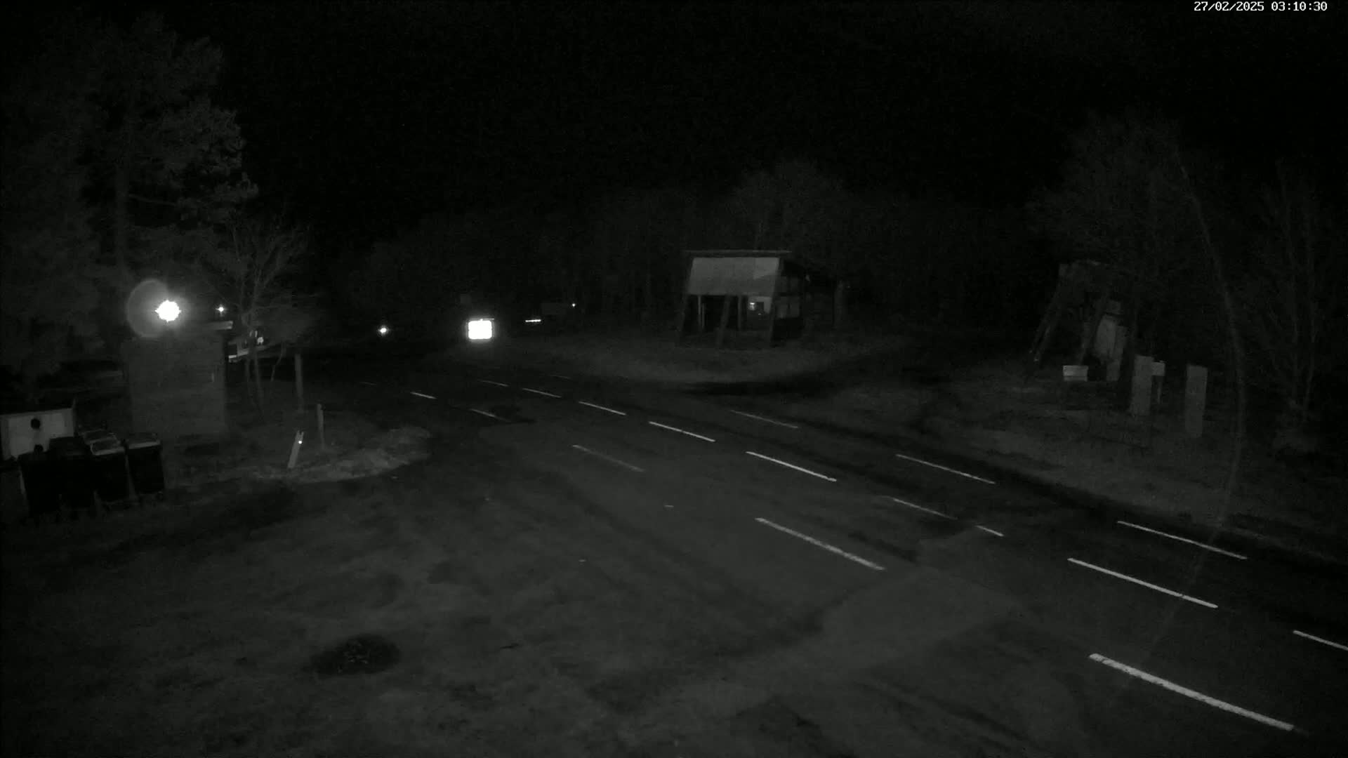 A nighttime, low-light view of a wet road with sparse, dark trees and a few structures in the background.