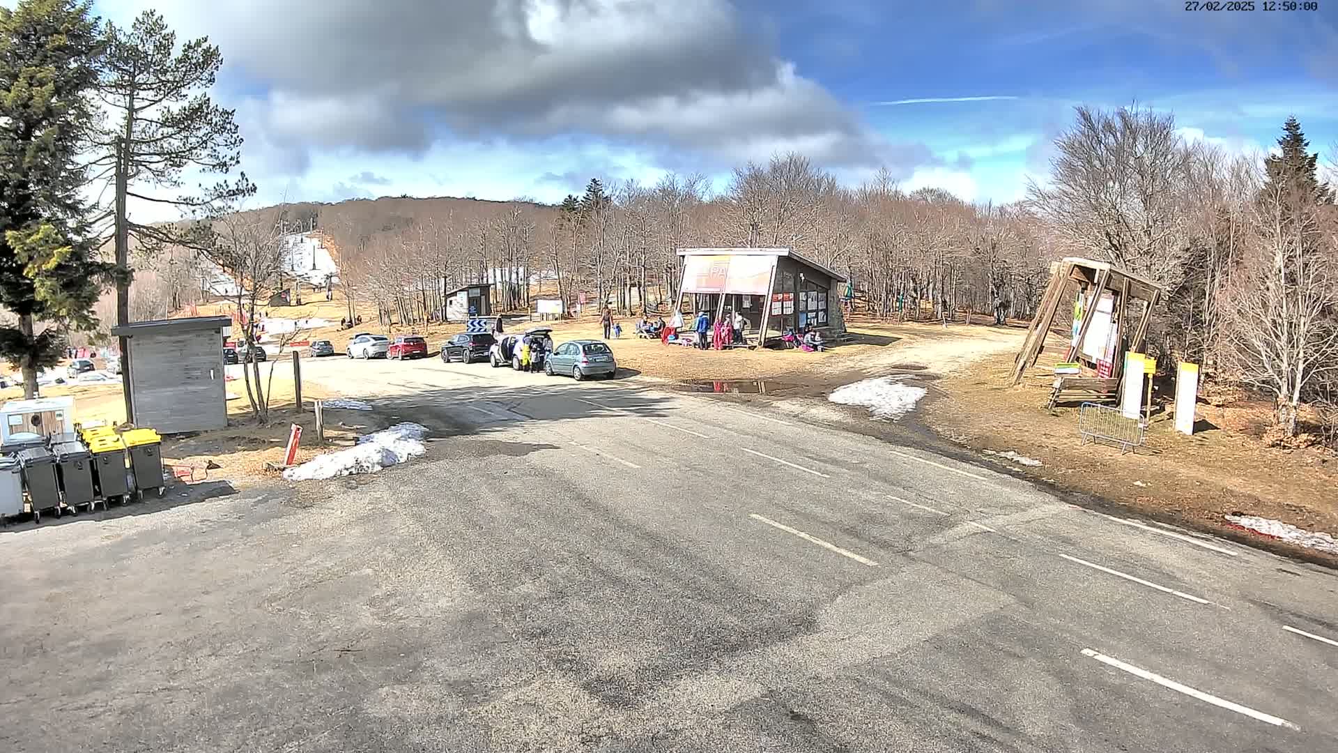 A paved road leads to a ski resort parking lot with several cars and a small building where people are gathered, under a partly cloudy sky.