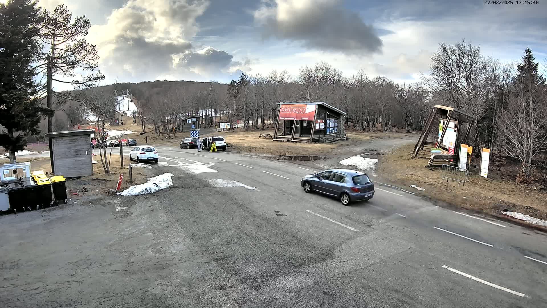 A mostly cloudy day at a ski resort parking lot with several cars, a small building, and some patchy snow on the ground.