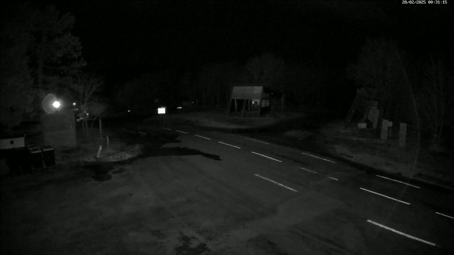 A dark, nighttime scene shows a mostly empty road intersecting with another road, surrounded by trees and a few structures under a clear, dark sky.