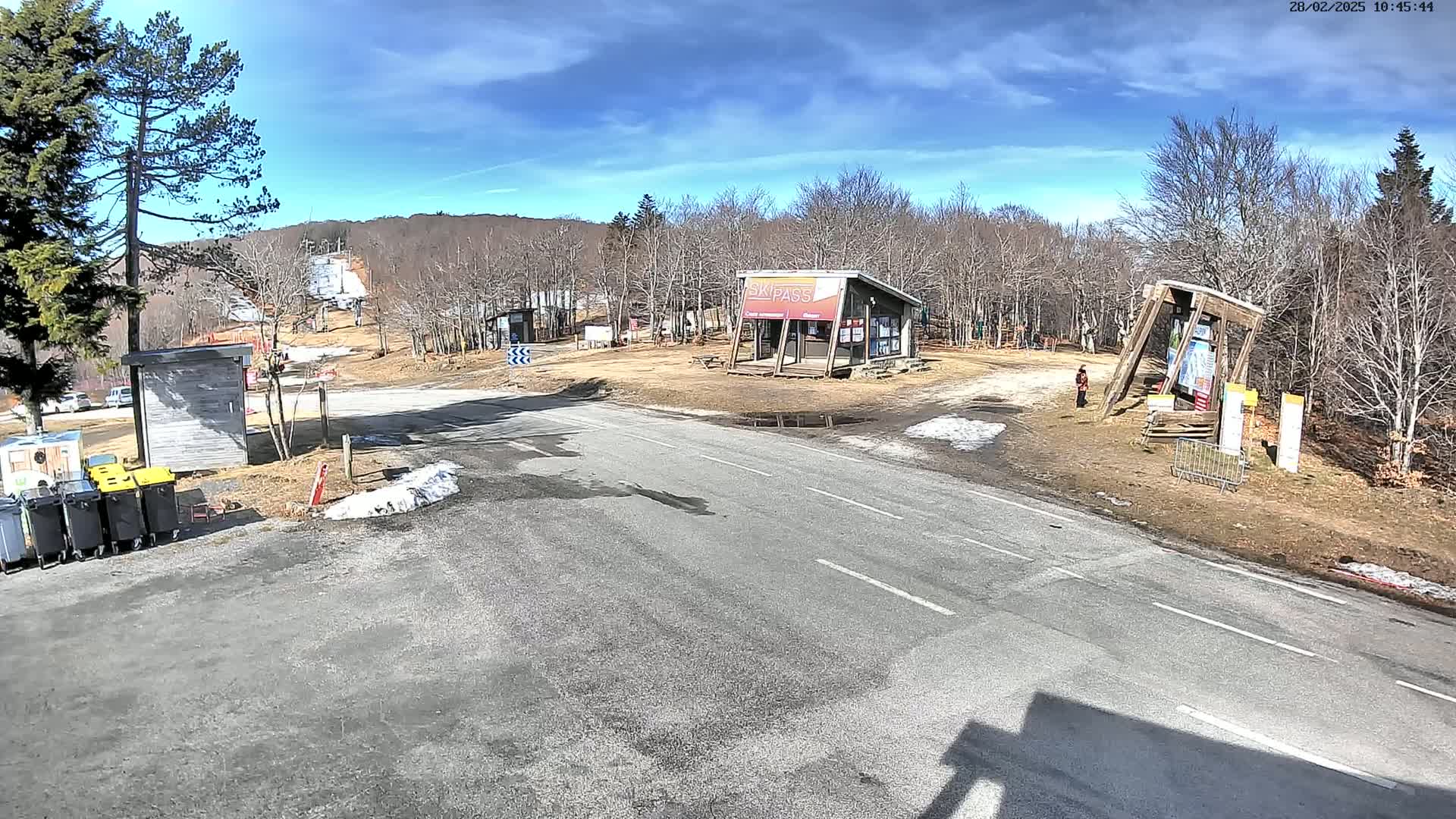 A paved road leads to a ski resort base area under a mostly sunny sky, with some small patches of snow and bare deciduous trees.