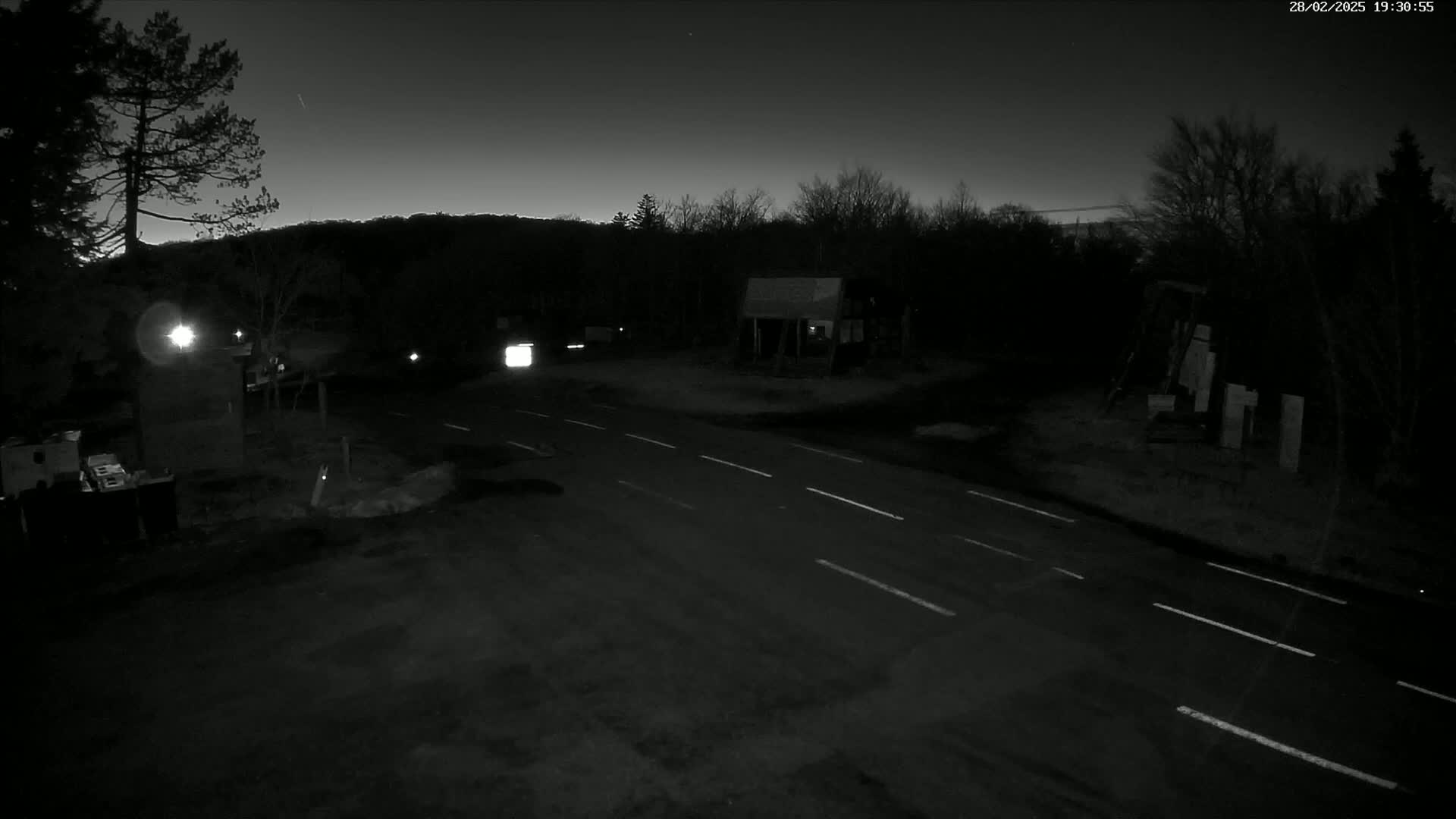 A dark, nighttime scene shows a two-lane road intersecting with another road, with a few trees and buildings visible under a clear, twilight sky.