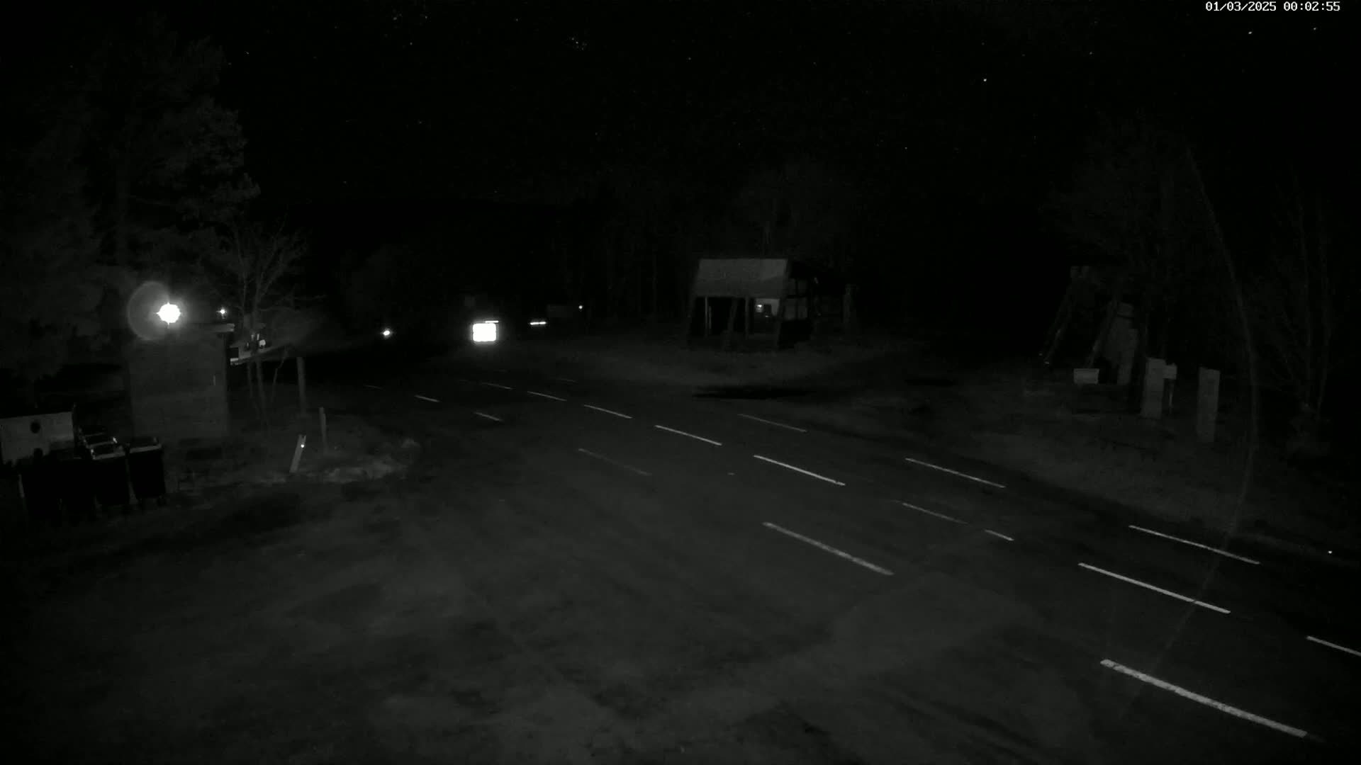 A dark, nighttime scene shows a road with faint white lane markings, a few vehicles with headlights, and some structures in the background under a starry sky.