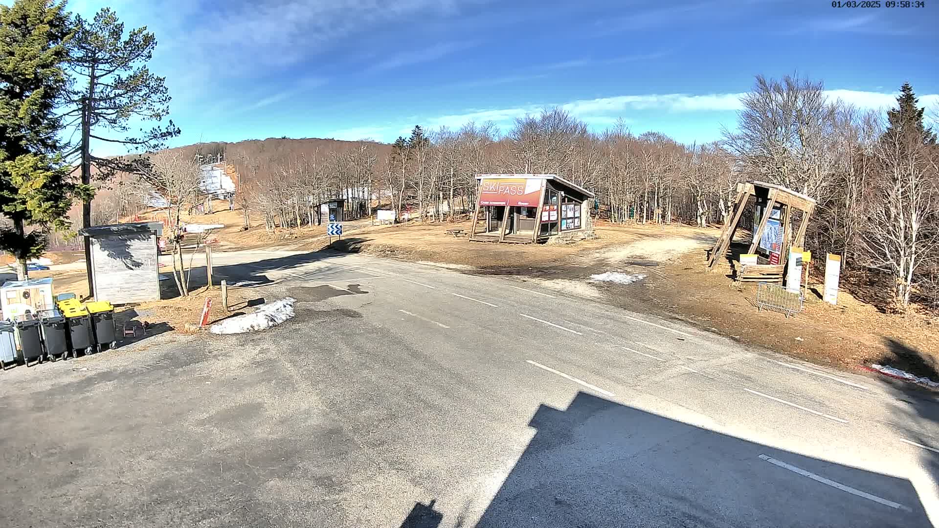 A sunny day at a ski resort parking lot shows a paved road leading to small buildings and a ski lift, with barren trees surrounding the area and patches of snow on the ground.