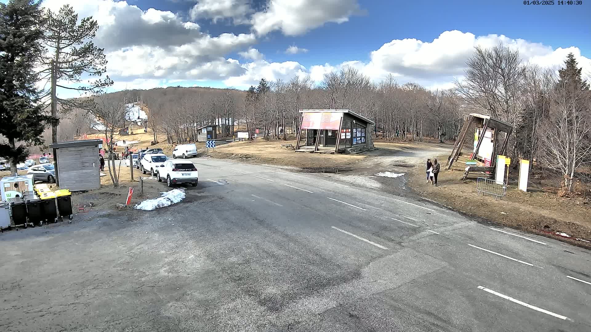 A paved road winds through a parking lot near a ski slope on a partly sunny day, with several cars parked and a few people standing near small buildings.