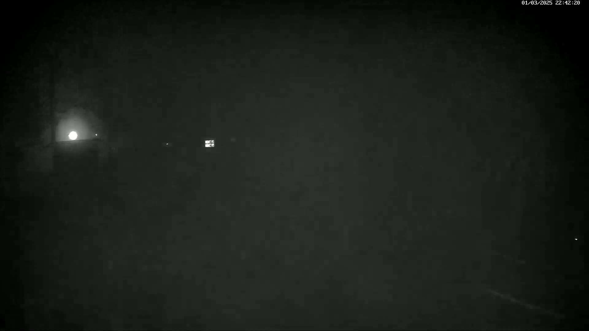 A nighttime, low-light view shows a dimly lit area with faint structures and a bright light source in the distance.