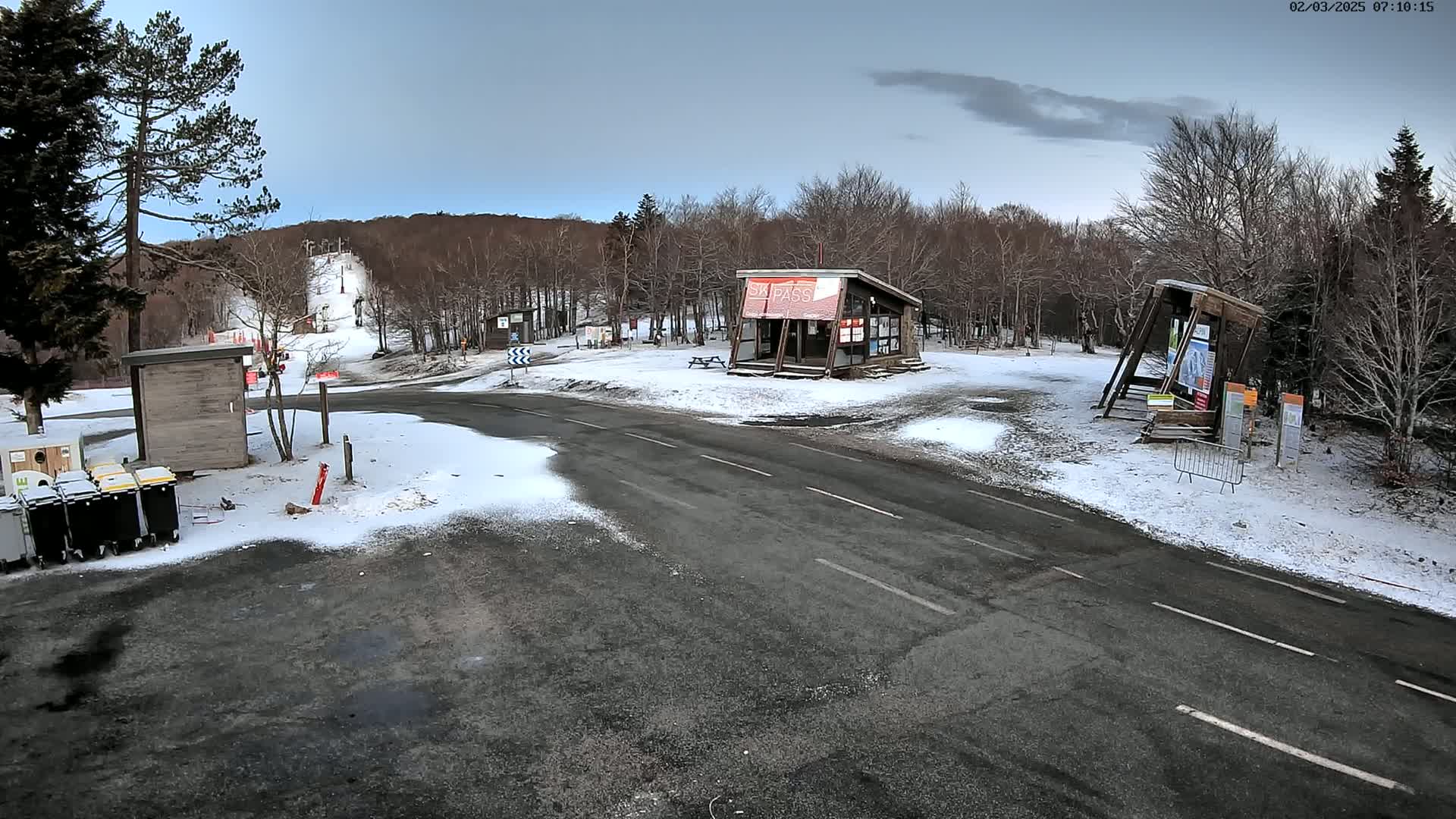 A snow-dusted parking lot and roadway are situated near a ski slope and several small buildings in a wooded area under a mostly cloudy sky.