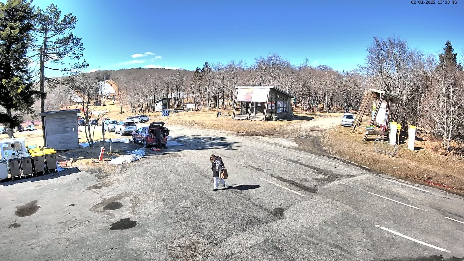 A woman walks across a paved road near a parking lot and several small buildings under a sunny sky in a sparsely wooded area.