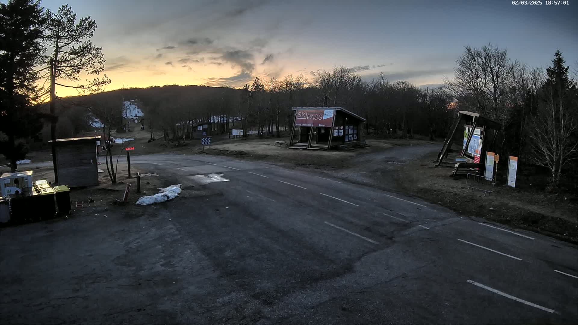 A paved road intersects at a ski resort parking lot during a dusky sunset, with some remaining snow on the ground and bare trees surrounding the area.