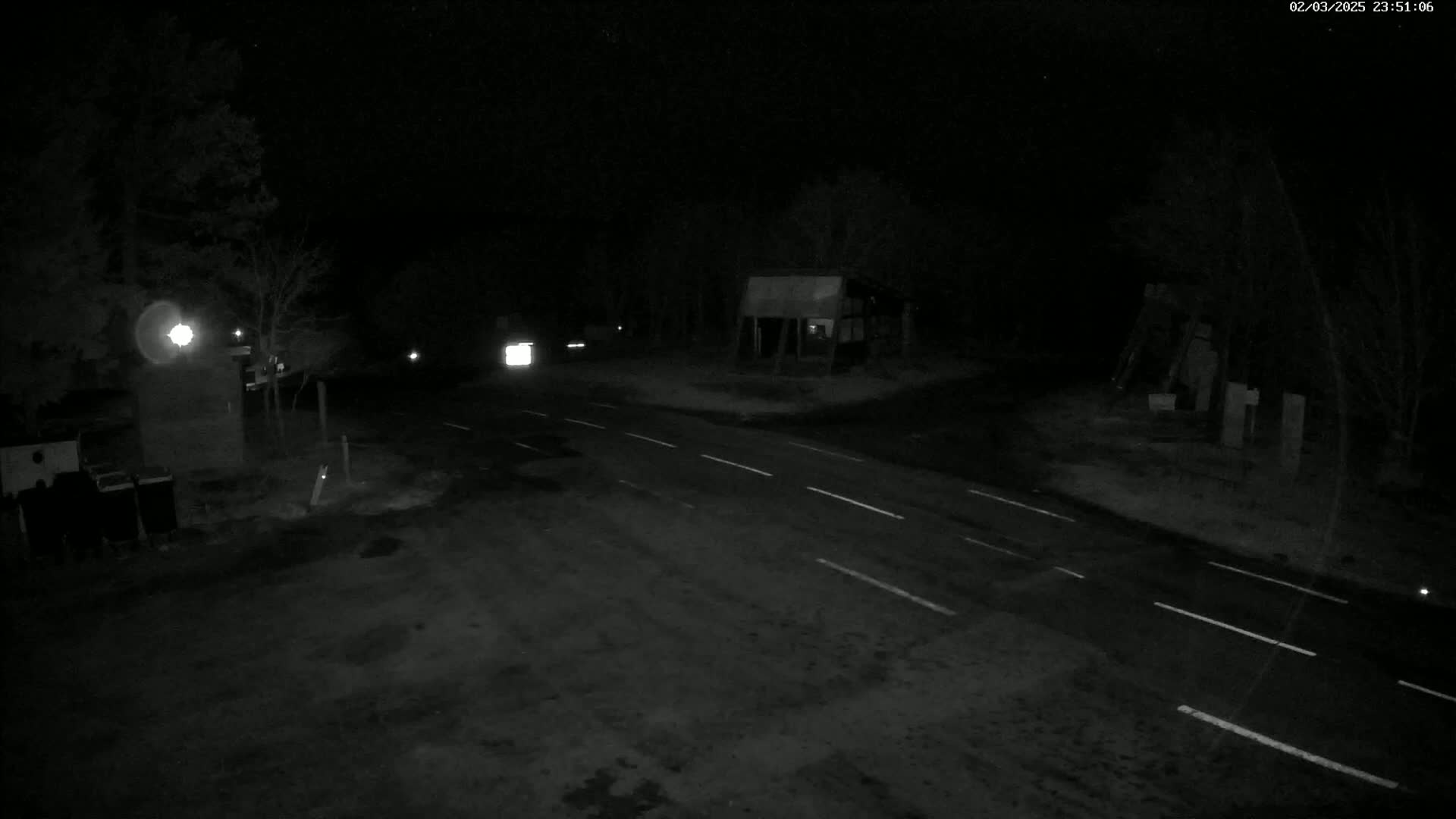A nighttime, low-light view of a dark road intersection with minimal traffic, surrounded by trees and structures.