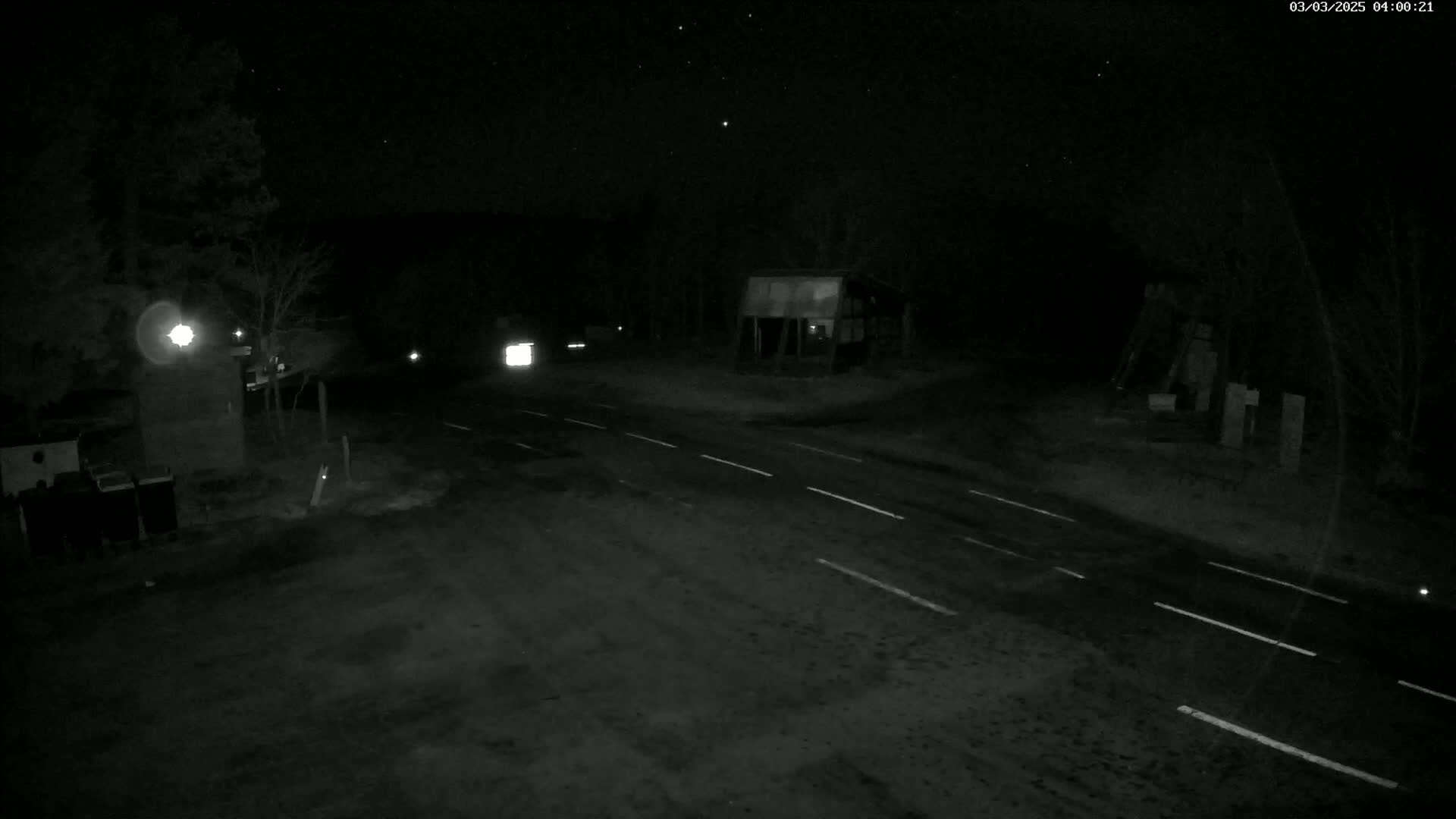 A dark, nighttime scene shows a road intersection with a few buildings and some vehicles, under a clear, starry sky.
