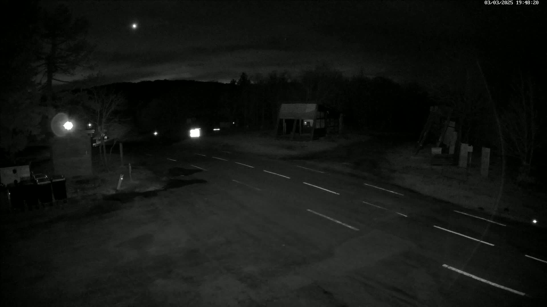 A nighttime, overhead view of a dark, empty two-lane road with a small structure and trees in the background under a partly cloudy sky.
