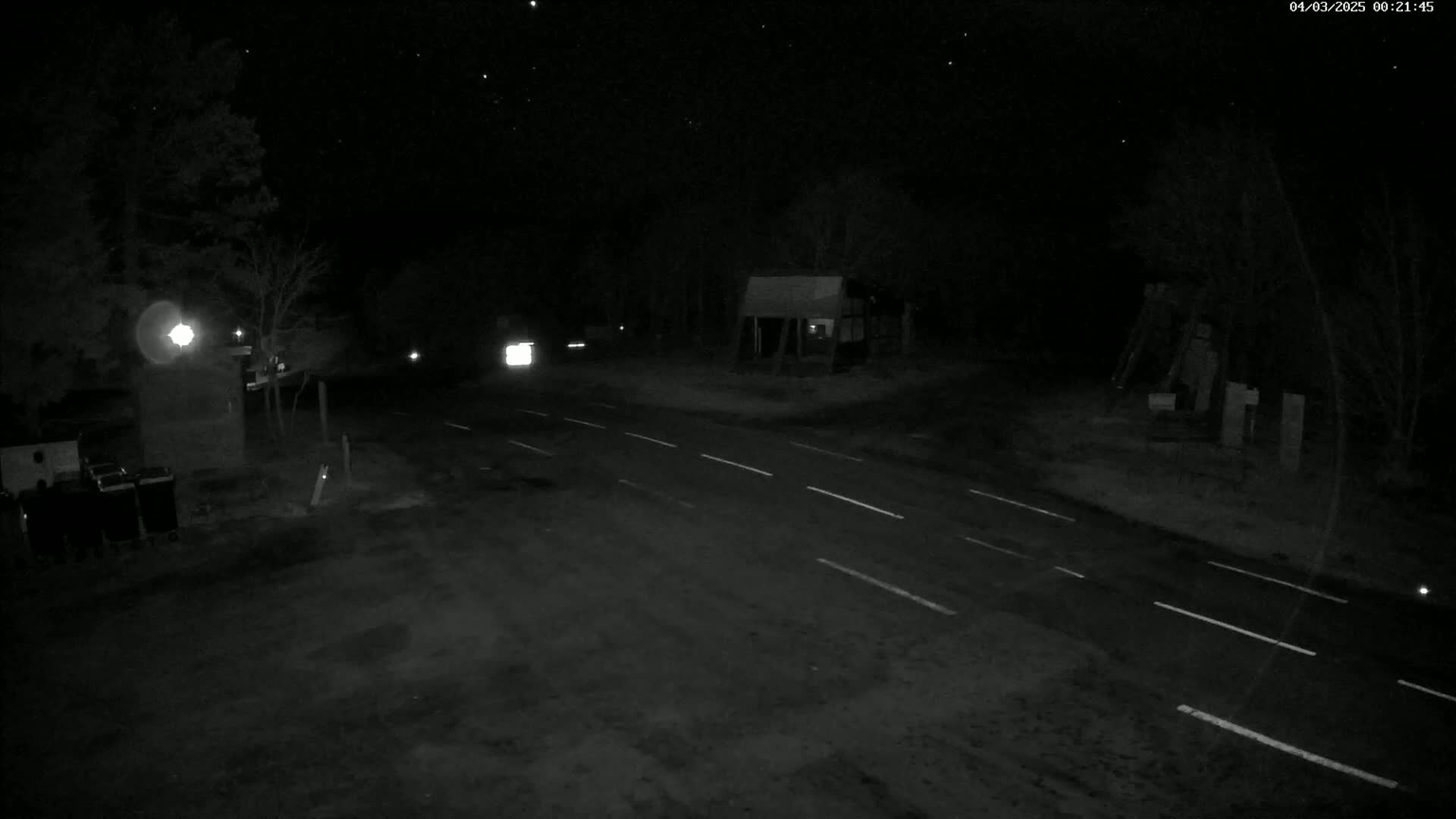 A dark, nighttime scene shows a sparsely-lit roadway intersection with a few vehicles and some structures visible under a clear, starry sky.