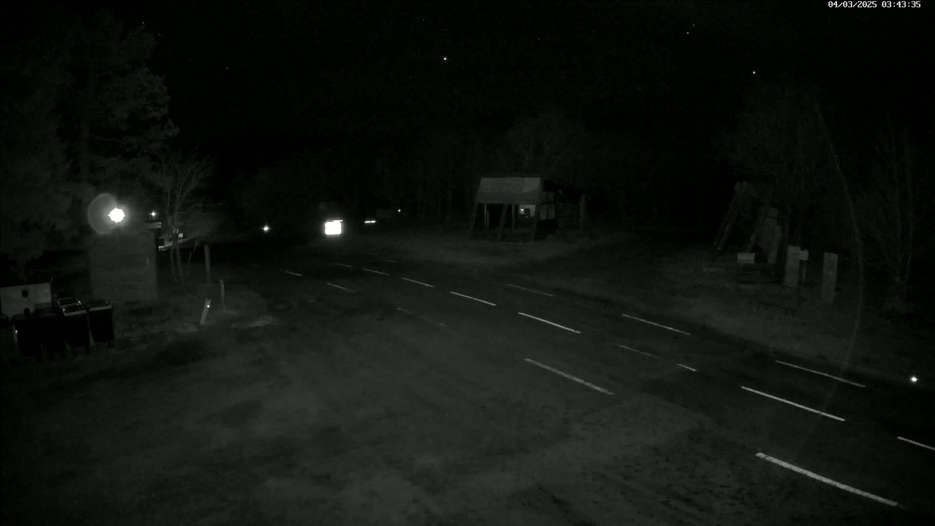 A dark, nighttime scene shows a road with white lane markings, sparsely lit by a single light source, with a few vehicles visible in the distance and trees lining the edges.