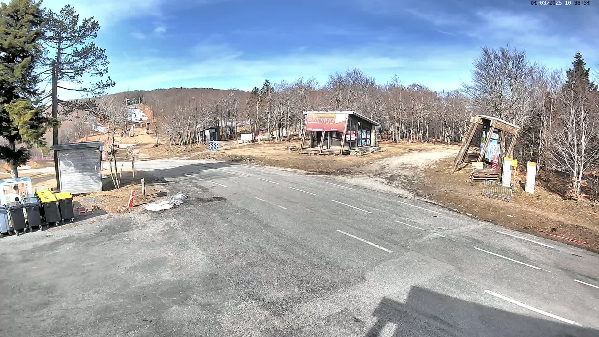 A paved road leads to a ski area under a mostly sunny sky, with bare deciduous trees and a few small buildings visible.