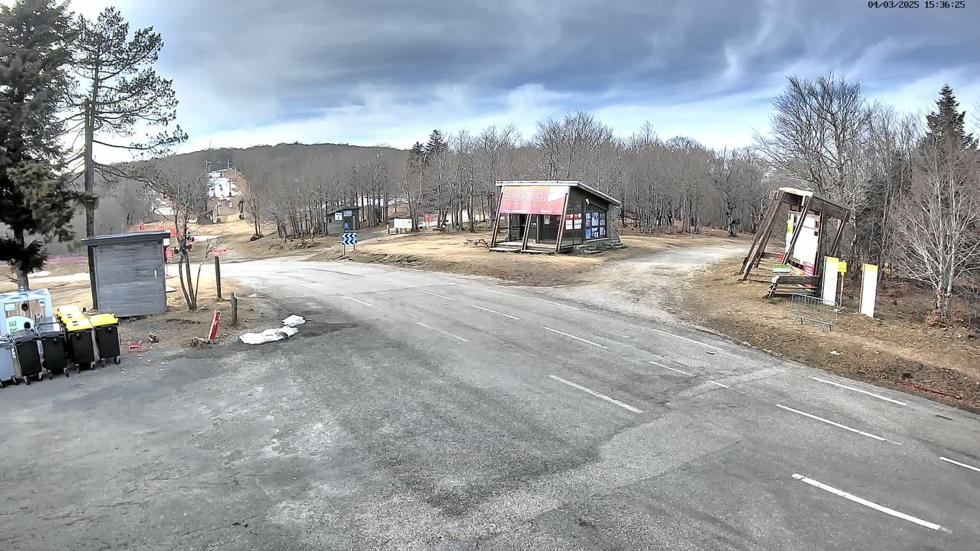 A paved road leads to a ski lift and several small buildings in a mostly barren, wooded area under a cloudy sky.