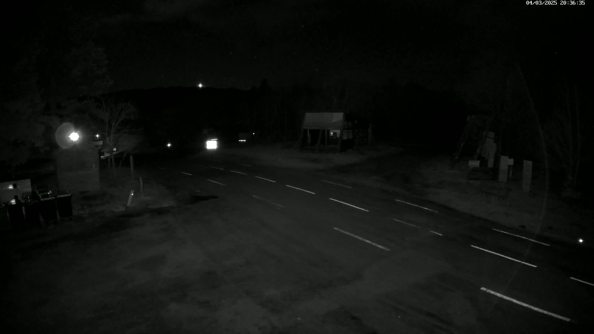 A nighttime view of a sparsely lit road intersection with a few vehicles and structures visible under a clear, starry sky.