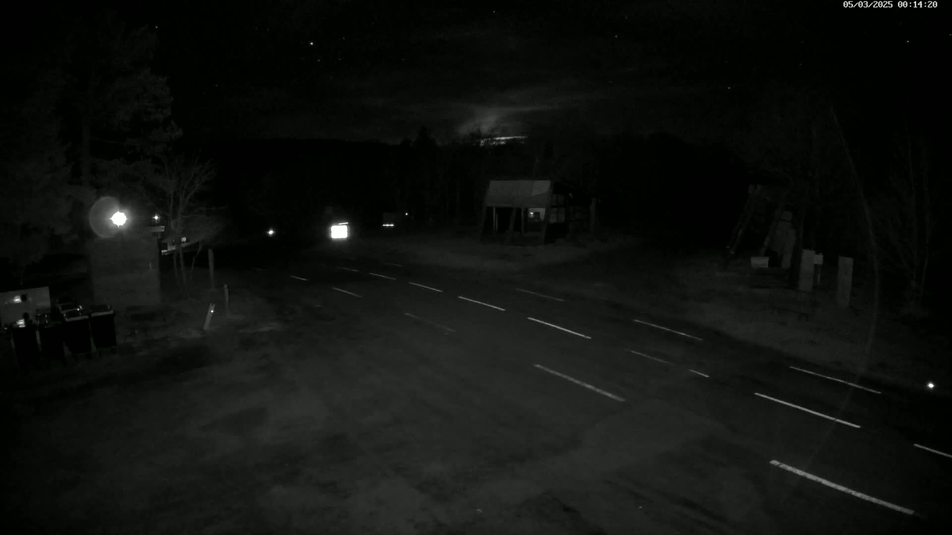 A nighttime view of a sparsely lit road intersection with a few vehicles and buildings visible under a partly cloudy sky.