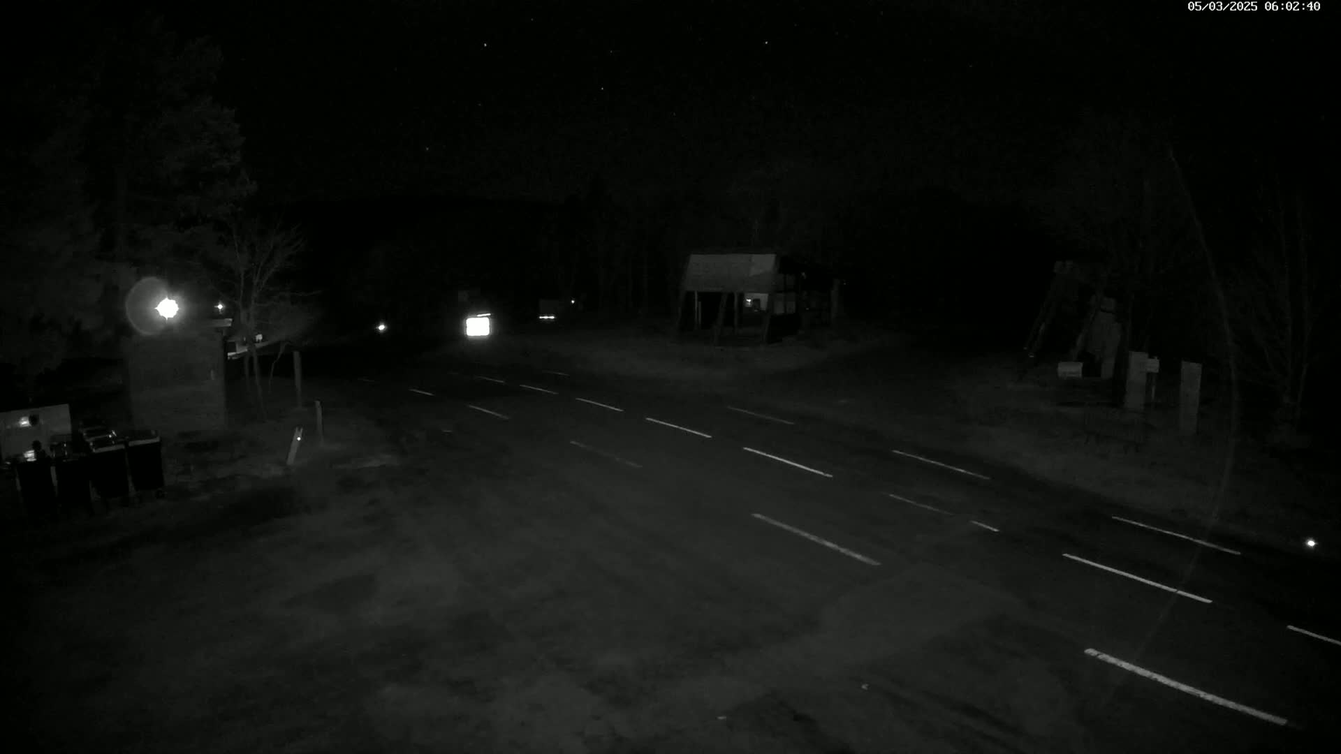 A dark, nighttime scene shows a sparsely lit road intersecting with another road, a few vehicles visible in the distance, under a clear, starlit sky.