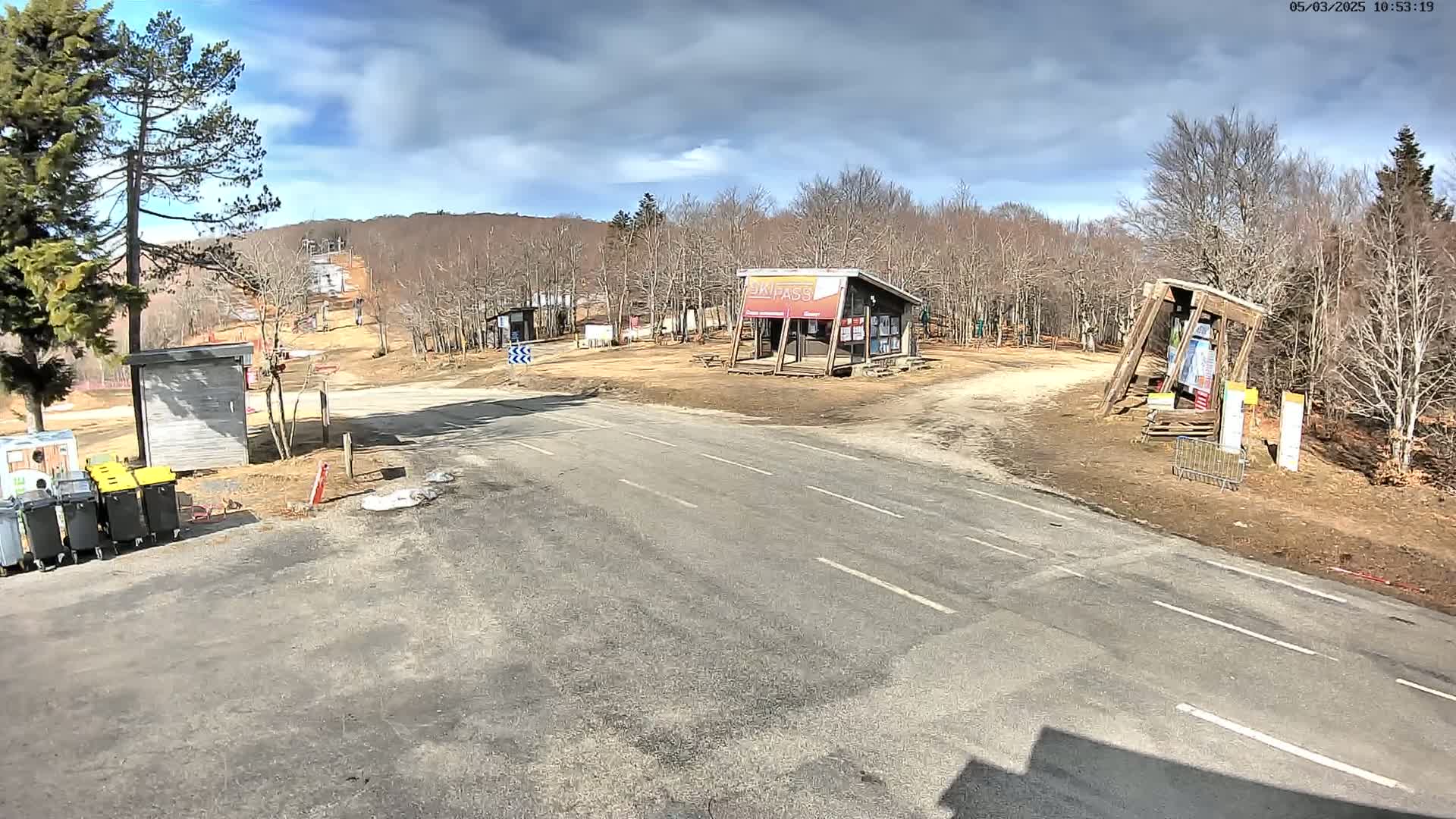 A mostly sunny day at a ski resort base area shows a paved road leading to several small buildings, a ski lift in the background, and leafless trees.