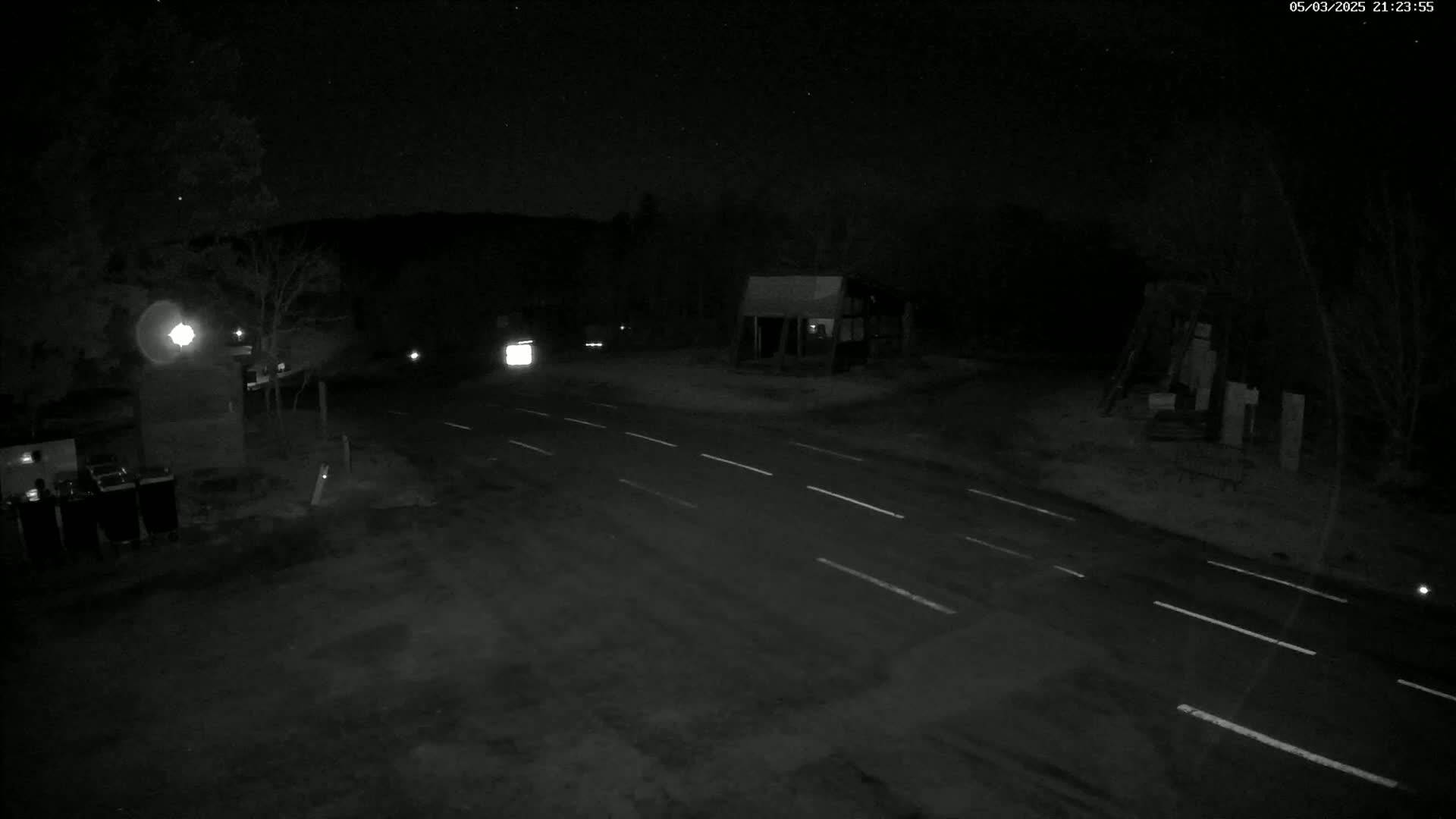 A dark, nighttime scene shows a sparsely trafficked two-lane road intersecting with another road, with a few vehicles visible in the distance under a clear, starry sky.