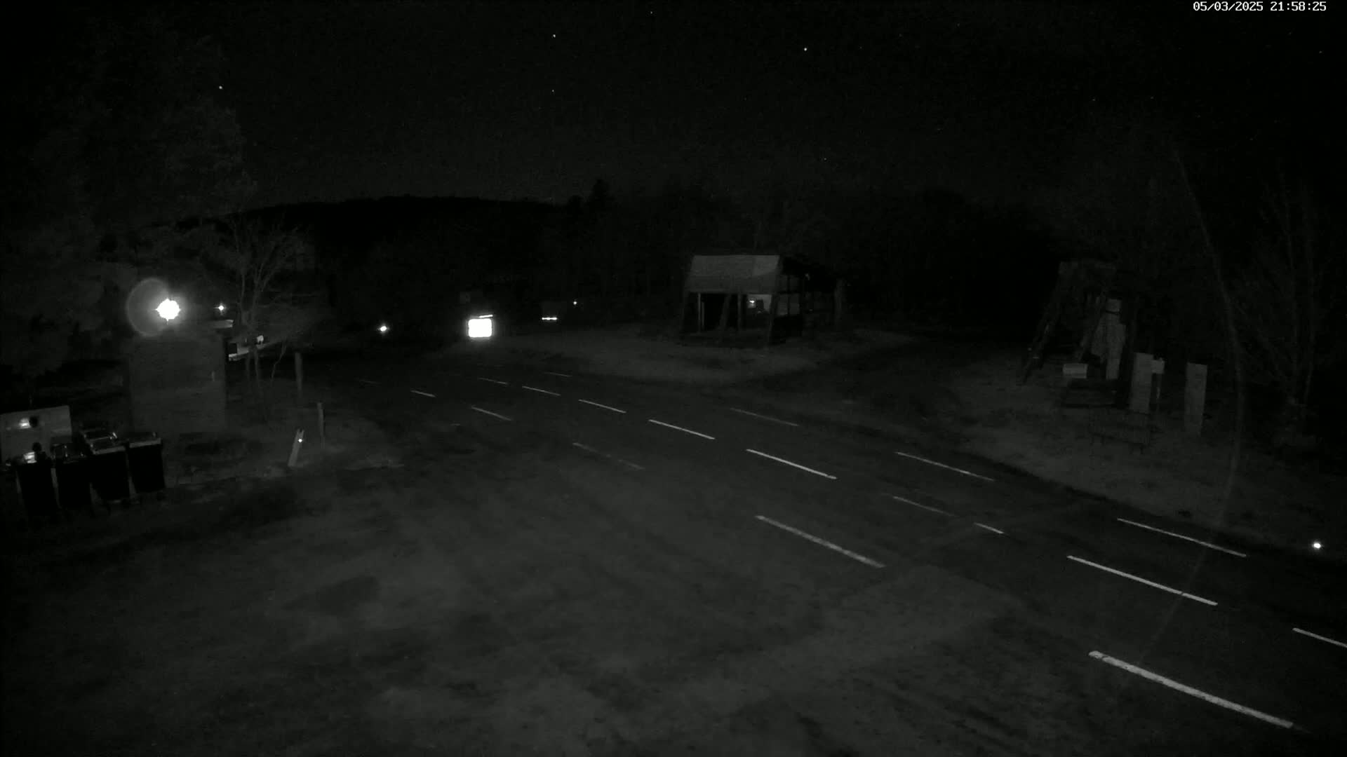 A dark, nighttime scene shows a road intersection with sparse vegetation and a small building under a clear, starry sky.