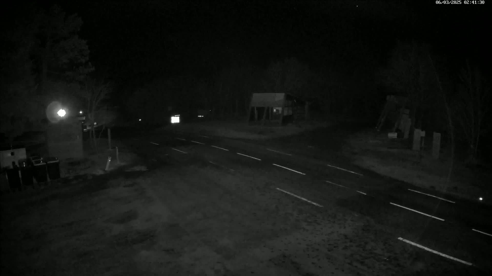 A nighttime view of a dark road intersection with a few indistinct structures and minimal vehicle traffic.