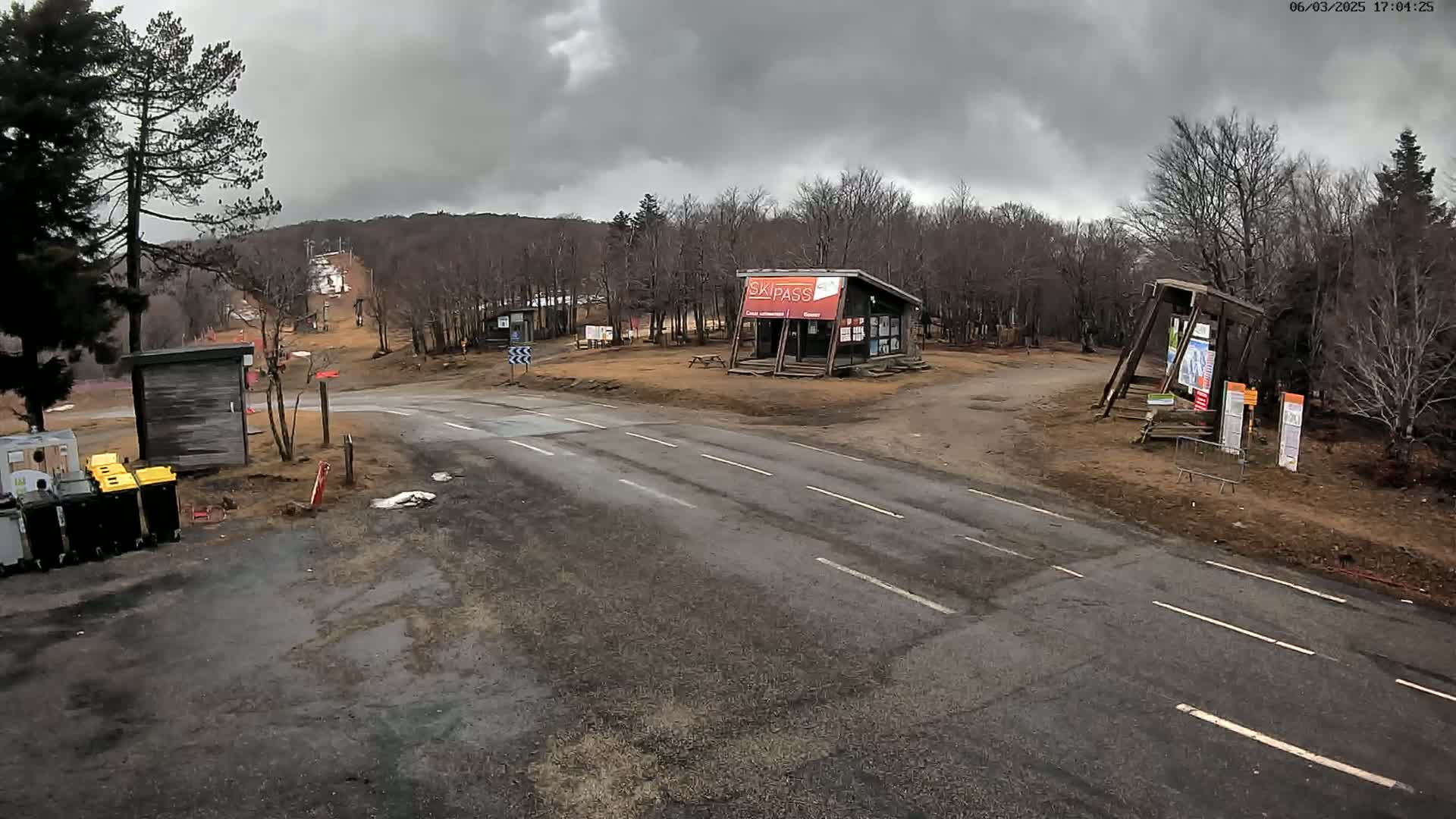 A paved road splits at a ski resort under a cloudy sky, with a small building and other structures visible, some of which appear to be informational kiosks.