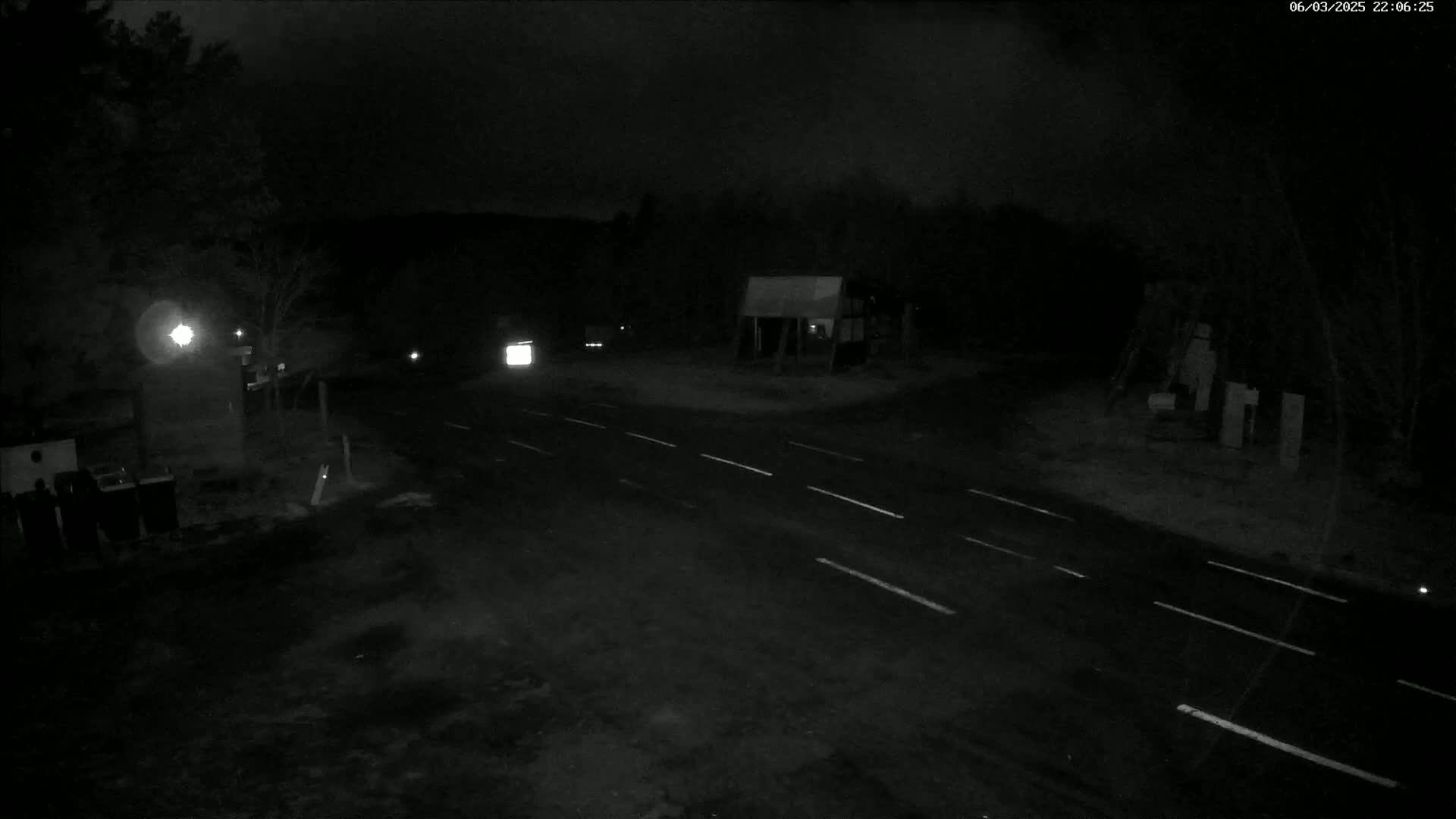A dark, nighttime scene shows a sparsely-lit road intersection with a few vehicles, trees, and small structures under a cloudy sky.