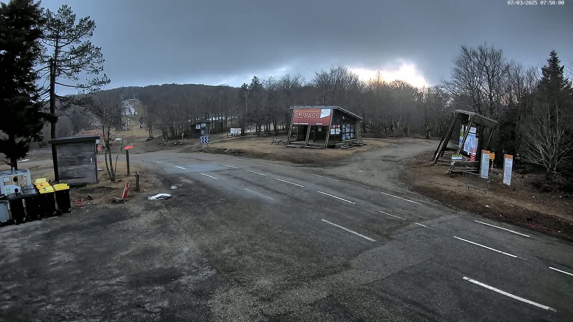 A paved road intersects with a dirt path in a wooded area under an overcast sky, with several small structures and a ski pass booth visible.
