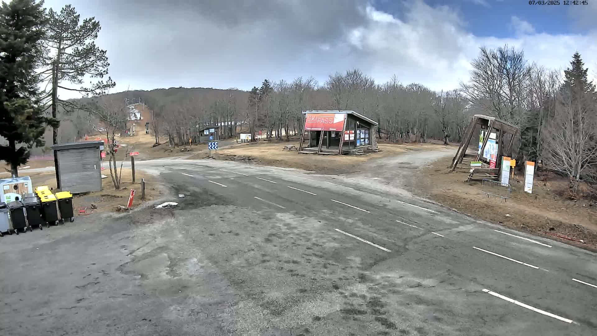 A paved road intersects with a dirt road at a ski resort under a partly cloudy sky, with bare trees and a ski lift visible in the background.