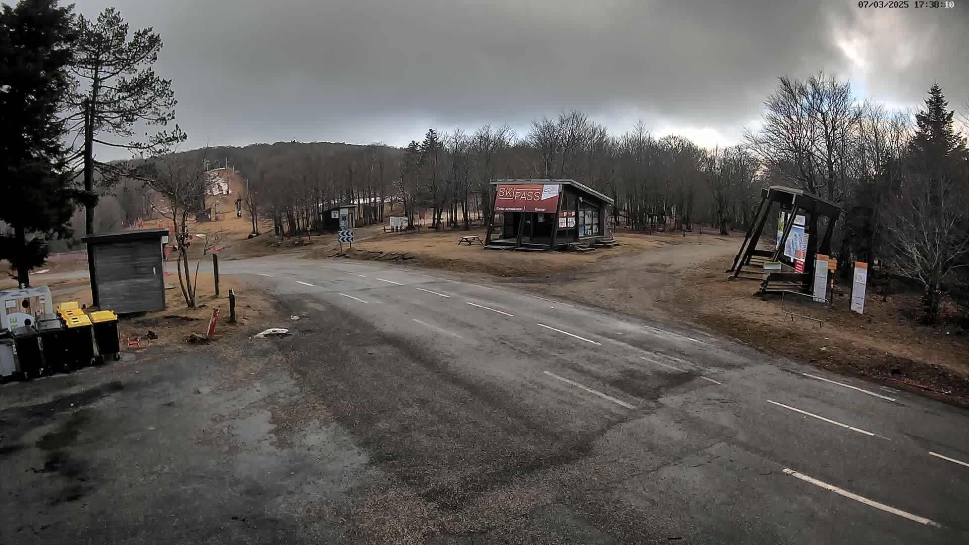 A paved road leads to a ski resort area under a cloudy sky, with several small buildings and a ski lift visible in the background.