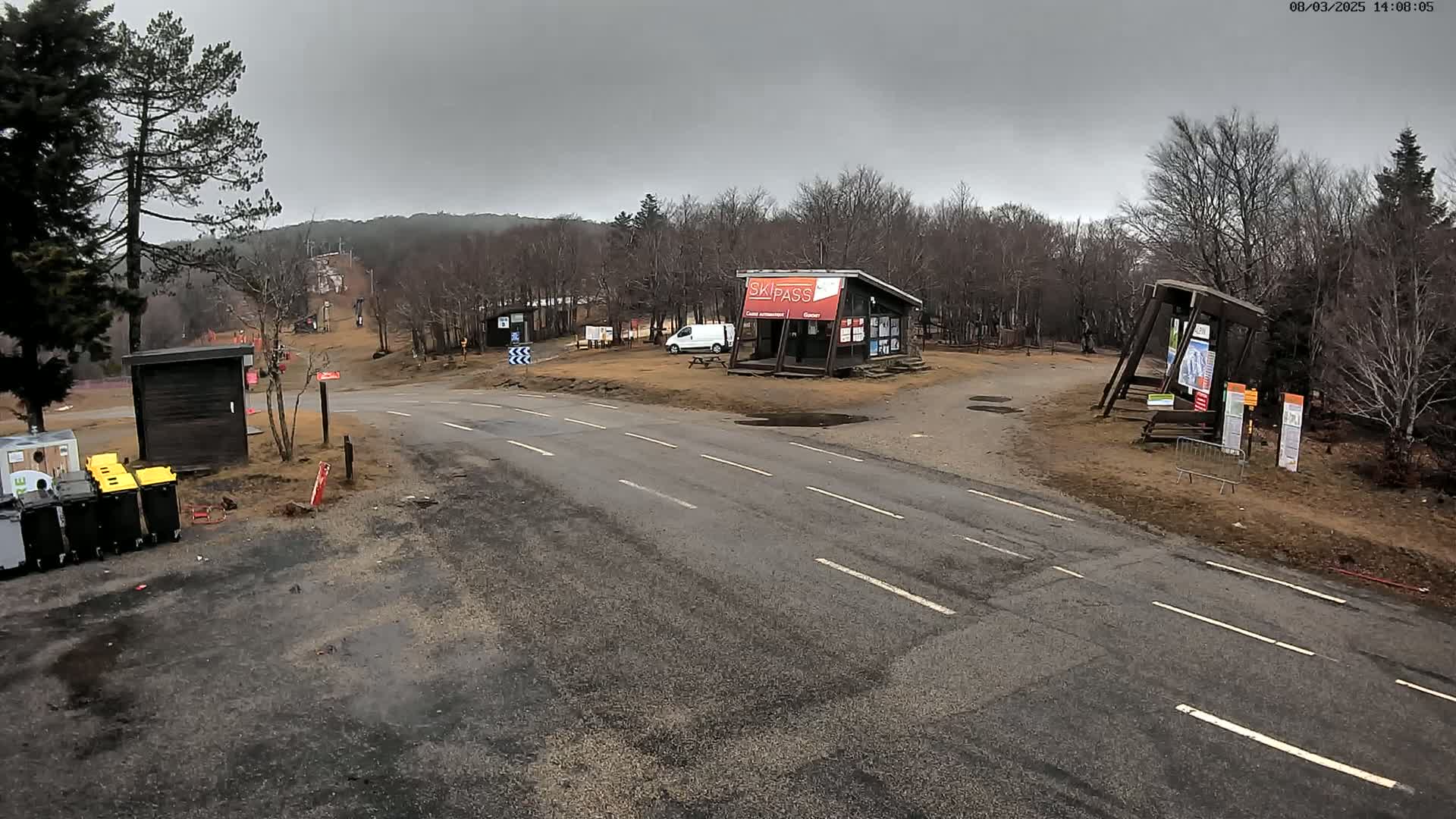 A paved road leads to a ski area with a small building, a van, and several informational signs under an overcast sky.