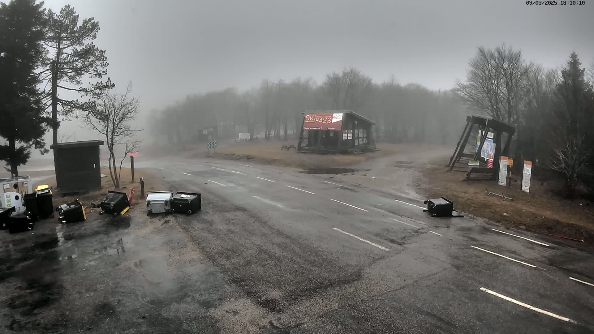 A foggy, wet road scene shows several overturned trash cans scattered near a small building and a parking area, with trees and other structures visible in the background.