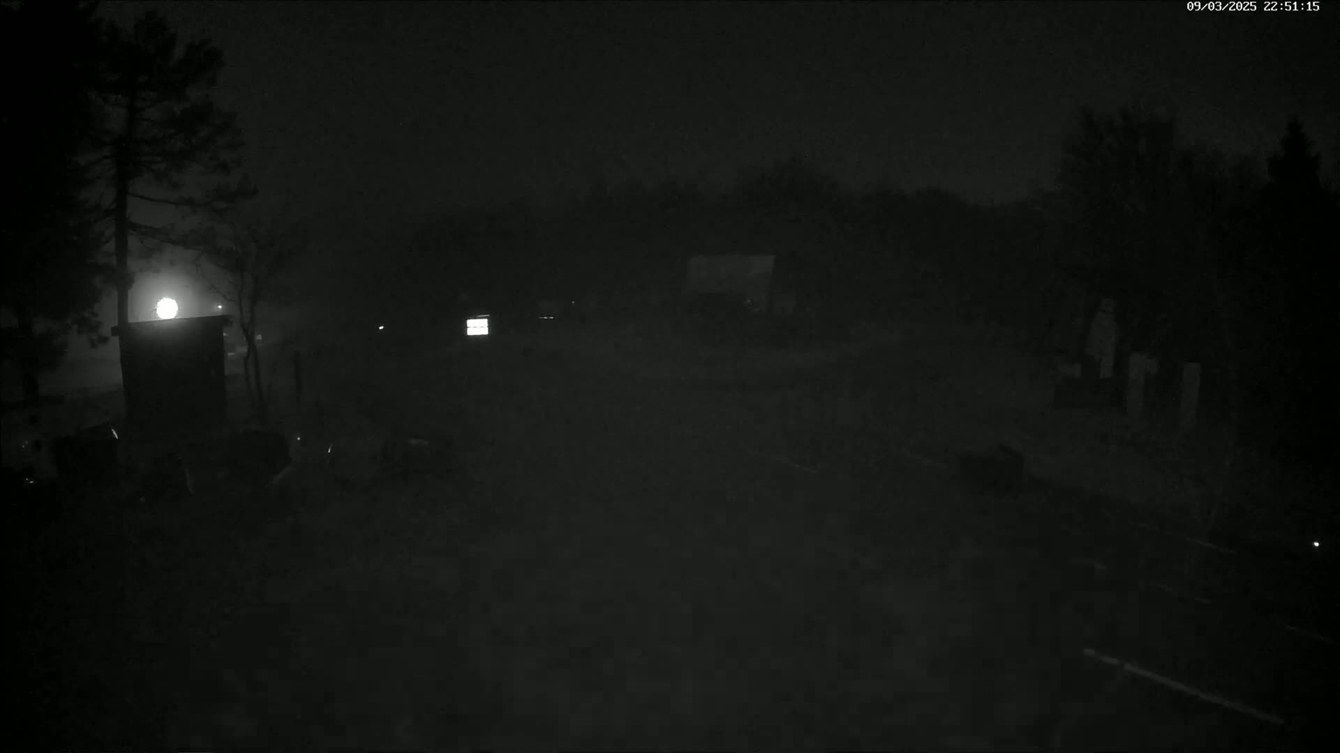 A dark, foggy nighttime scene shows a sparsely lit area with trees, some structures, and what appear to be vehicles.
