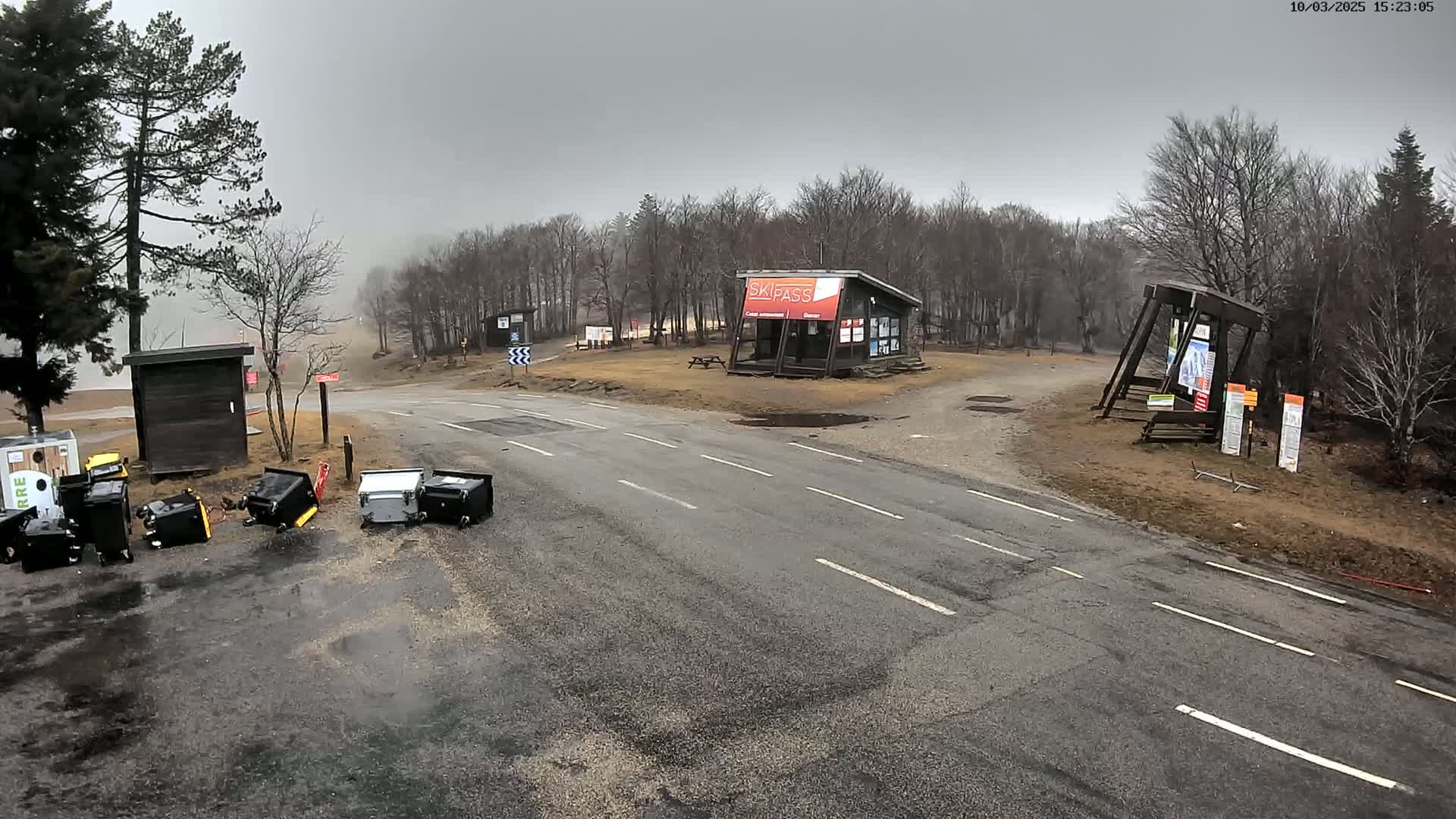 A paved road intersects with a dirt road in a misty, outdoor setting, where several overturned trash cans litter the ground near a small building.