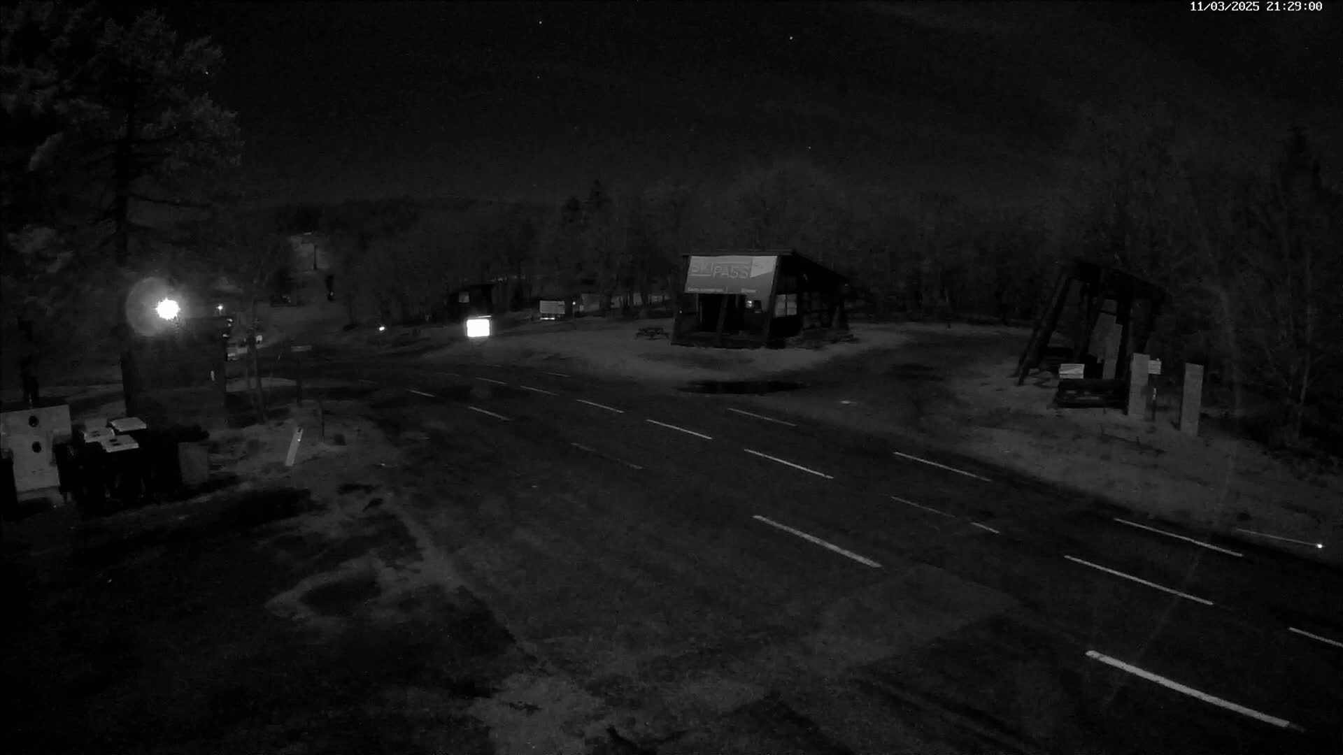 A nighttime, snow-covered roadway intersects with a small parking area containing a few buildings under a dark, starry sky.