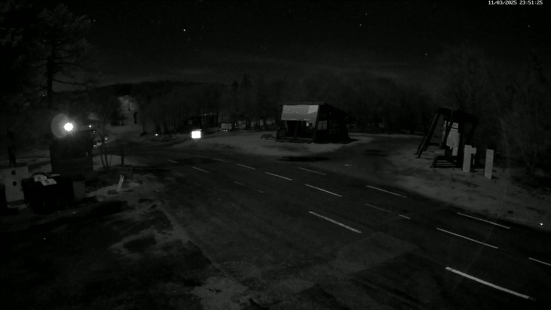 A nighttime, snowy scene shows a road intersection with a small building and surrounding trees under a starry sky.