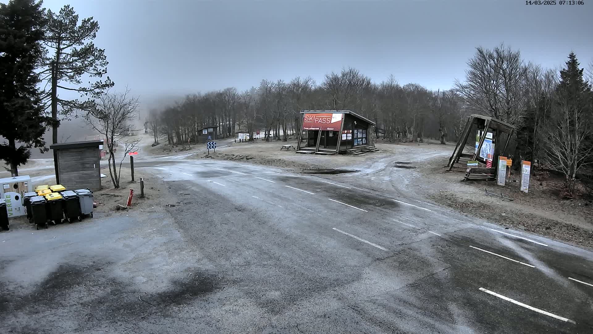 A paved road intersection at a ski resort on an overcast day, with small buildings and trash receptacles nearby, surrounded by leafless trees.