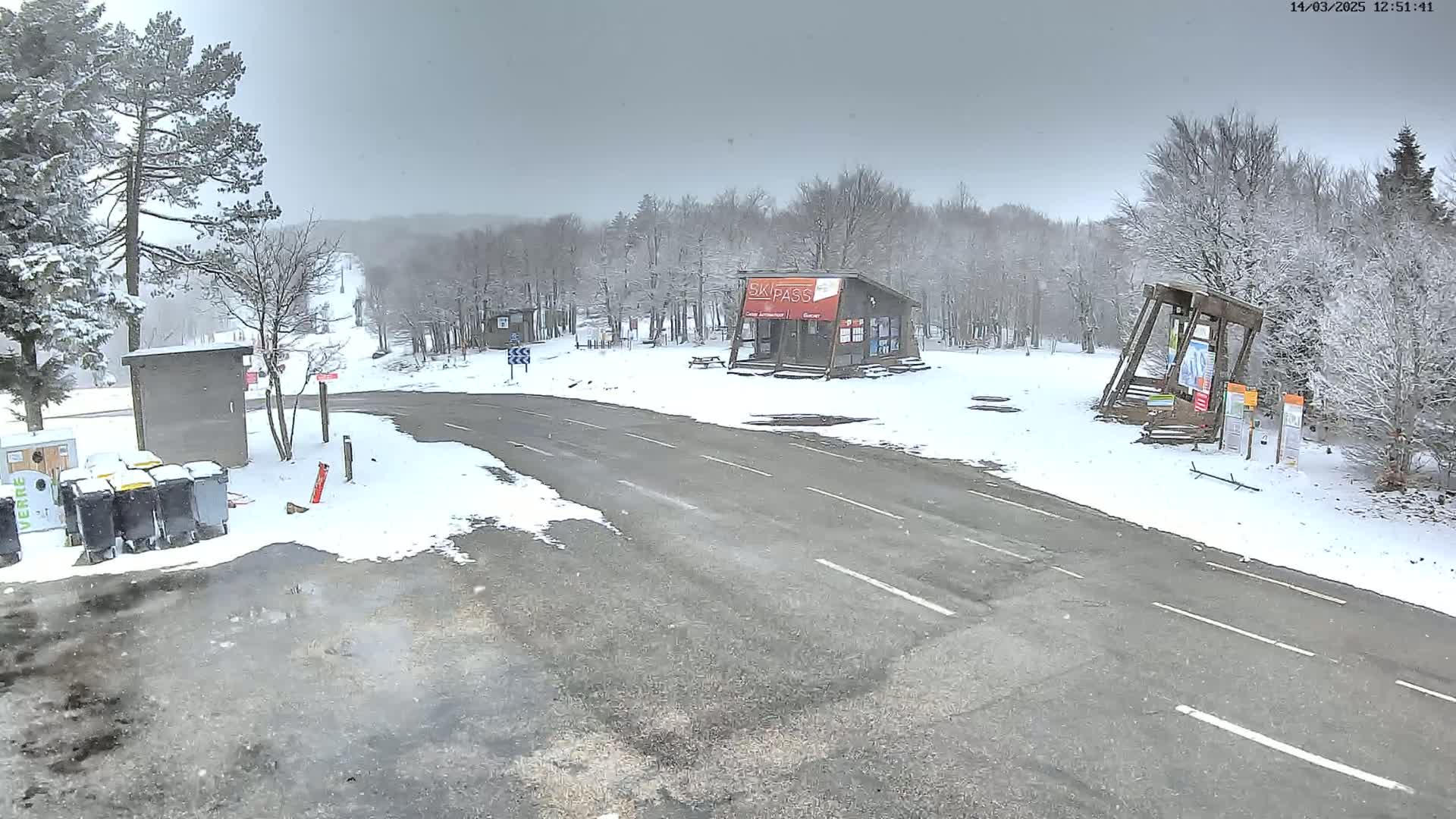 A snow-covered parking lot and road are seen next to a small building and a ski lift base, with a snowy forest in the background under an overcast sky.