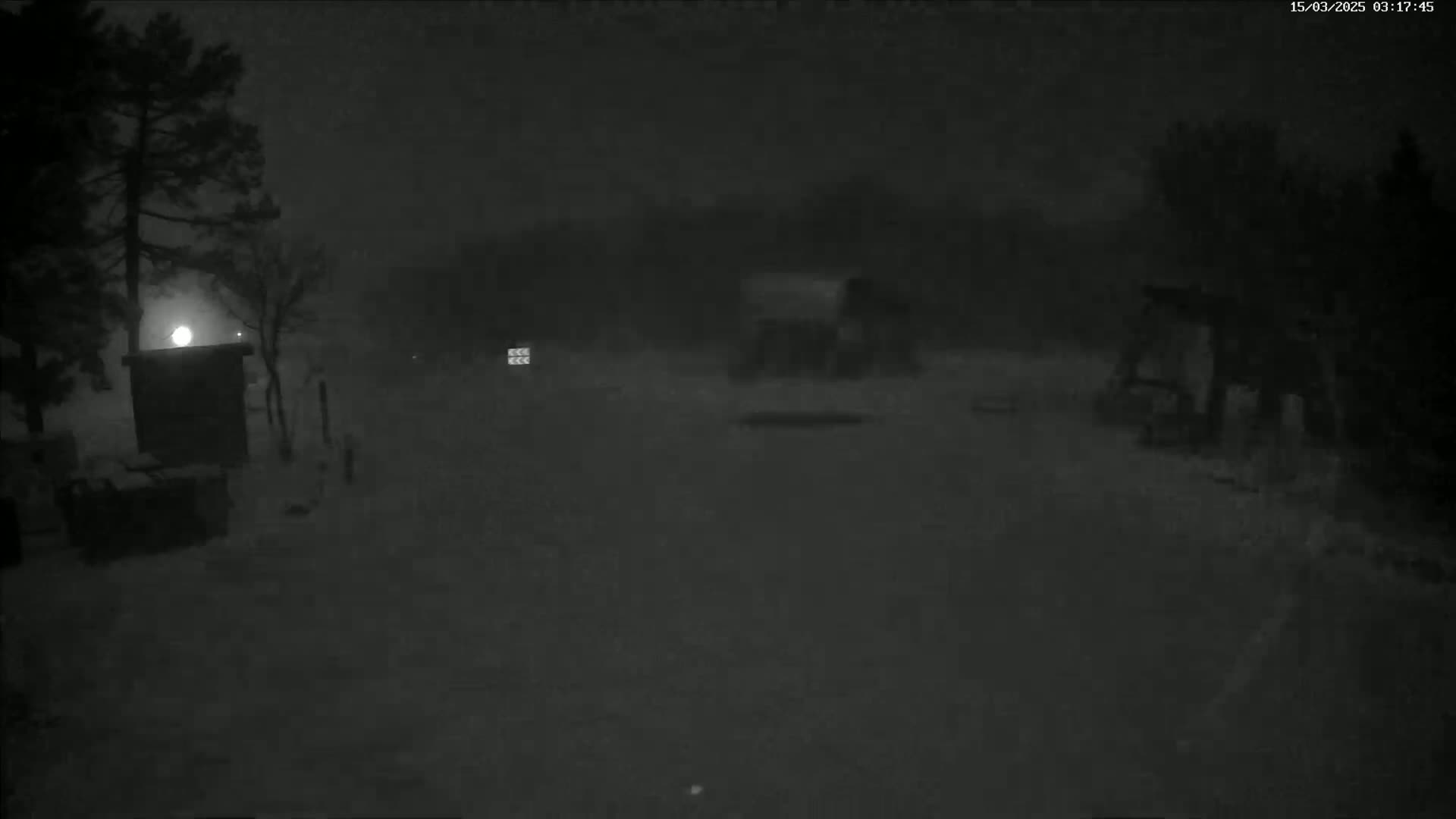 A snow-covered, dimly lit area at night, showing a small structure and some trees under heavy snowfall.