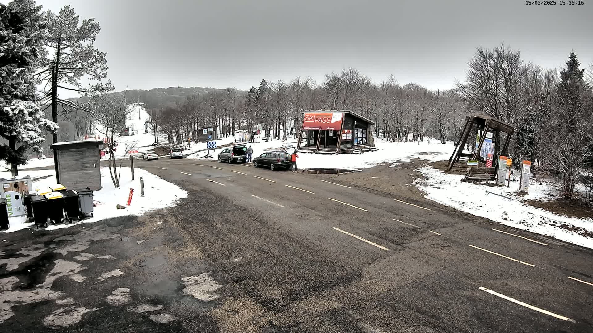 A snowy parking lot with several cars and a ski pass building is next to a snow-covered road leading to a ski slope in a forest under a cloudy sky.