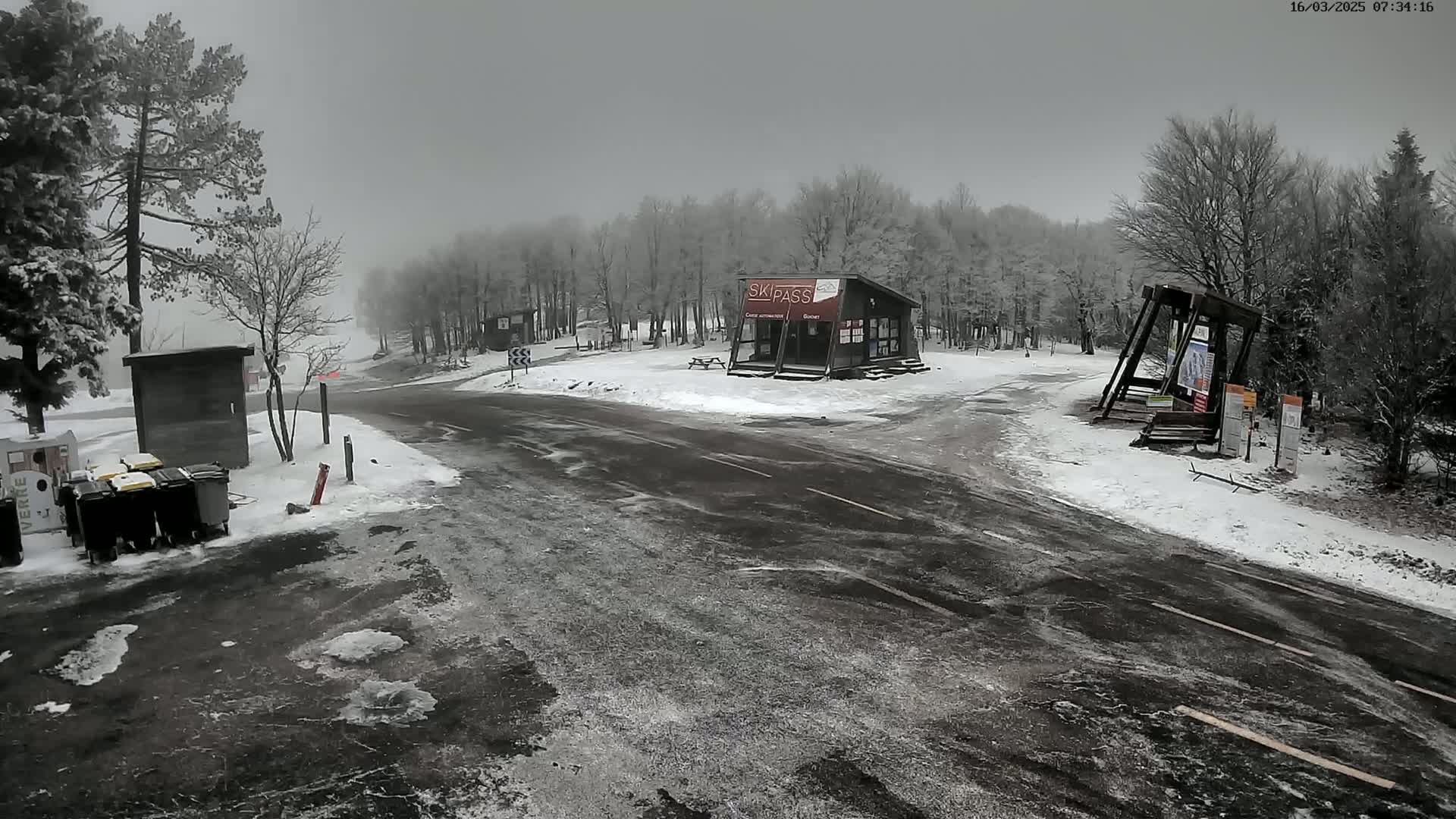 A snow-covered parking lot and road are seen in front of a small ski pass building, with leafless trees and overcast skies in the background.