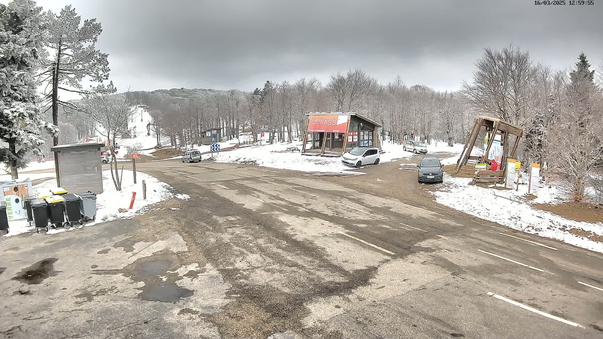 A snowy parking lot and roadway at a ski resort under an overcast sky shows several cars and a ski pass building.